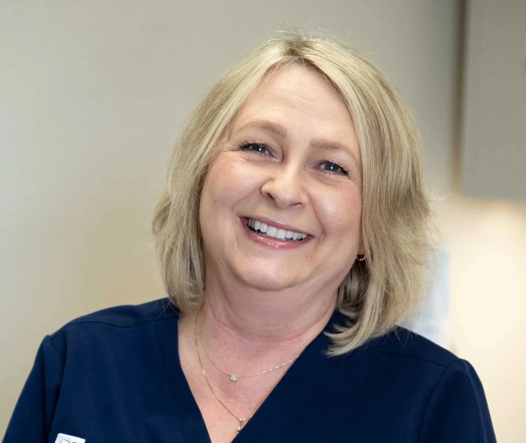 Smiling middle-aged woman with shoulder-length blonde hair, wearing a navy blue top and jewelry, in an indoor setting.