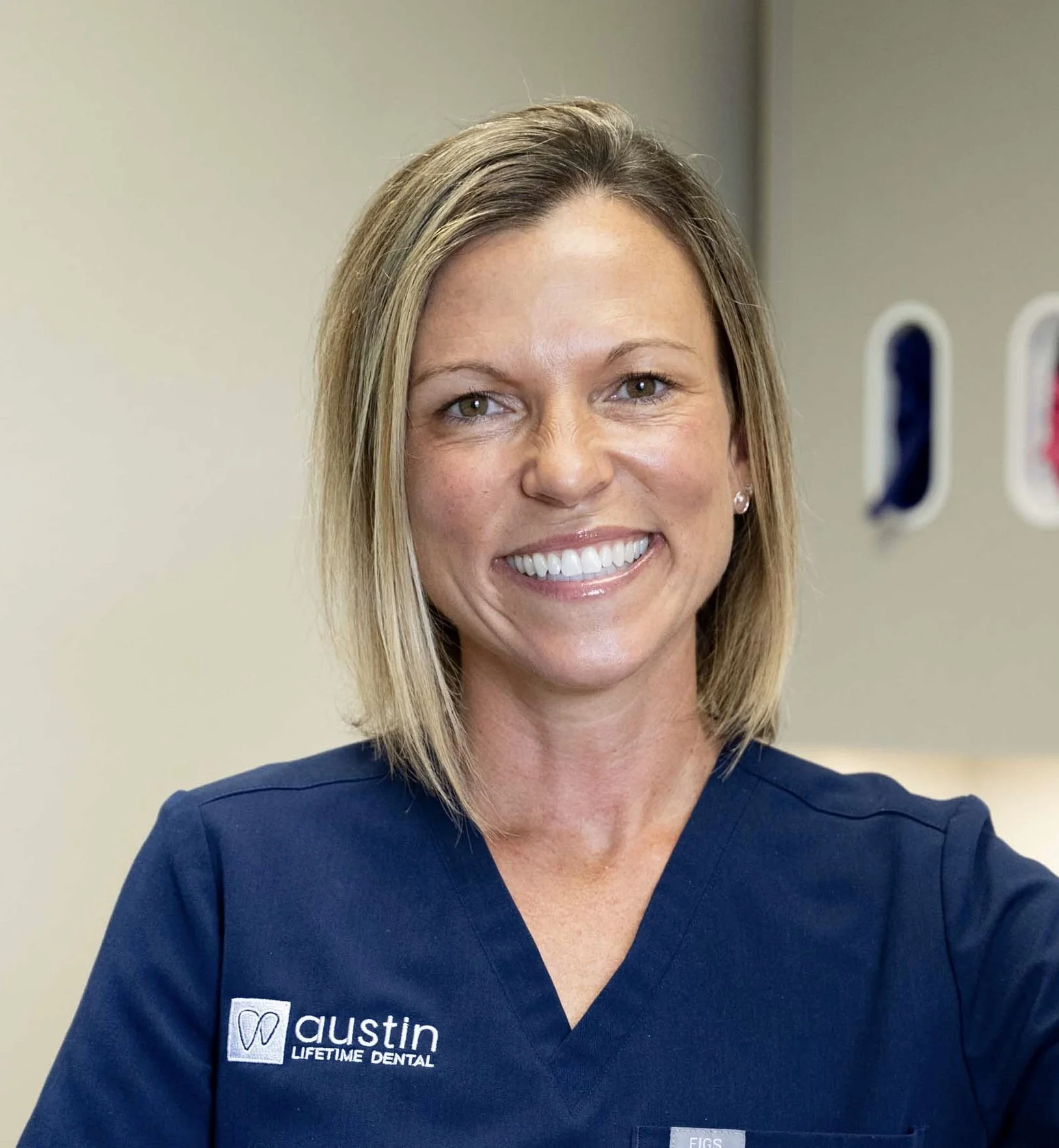 A woman smiling in a navy blue dental uniform with the logo and text 'Austin Lifetime Dental' on it, standing in a dental office.