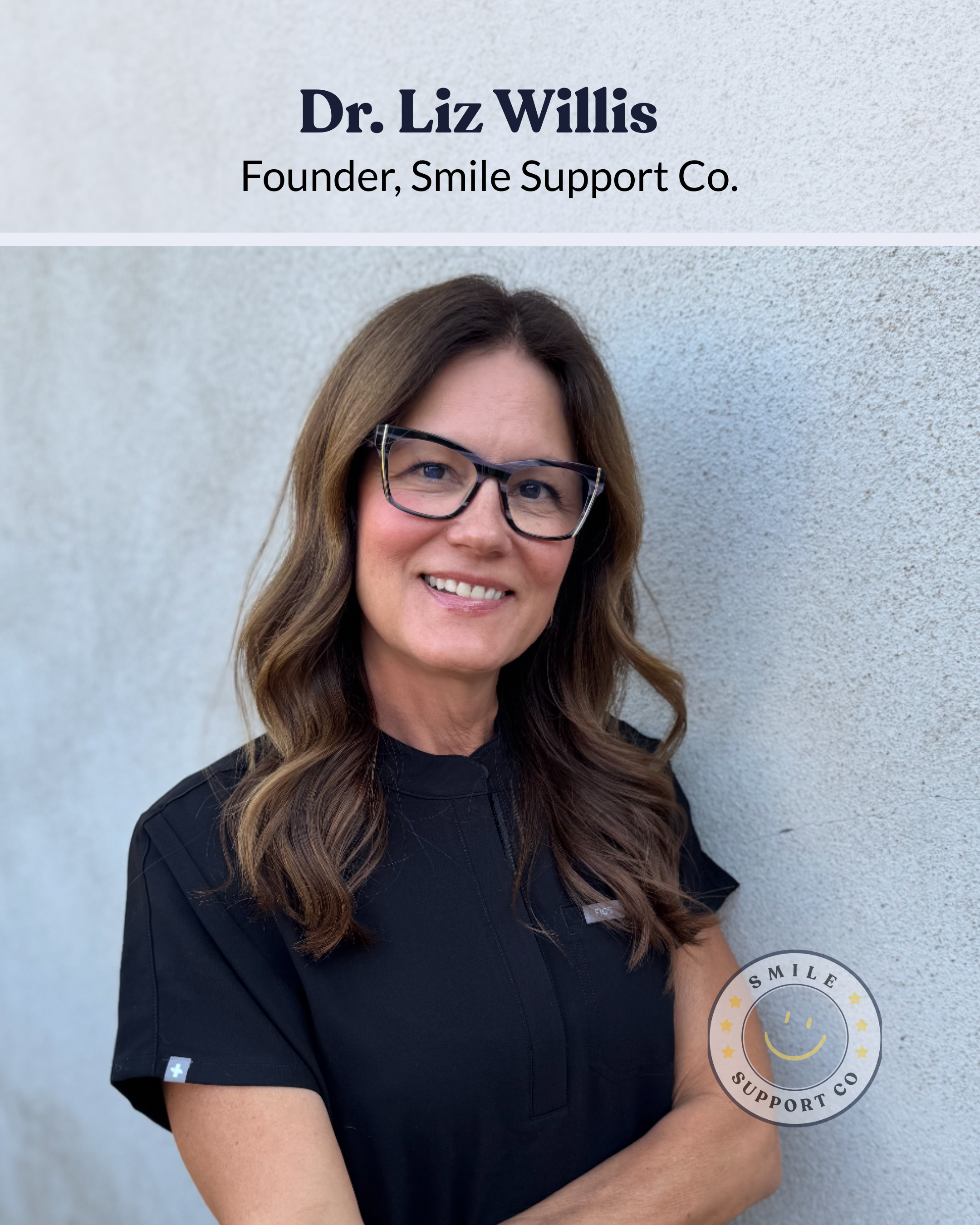 Portrait of Dr. Liz Willis, Founder of Smile Support Co., standing against a light-colored wall, smiling, wearing glasses and a black scrub top.