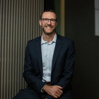 A smiling man in a navy suit with glasses, white shirt, sitting in front of a dark background