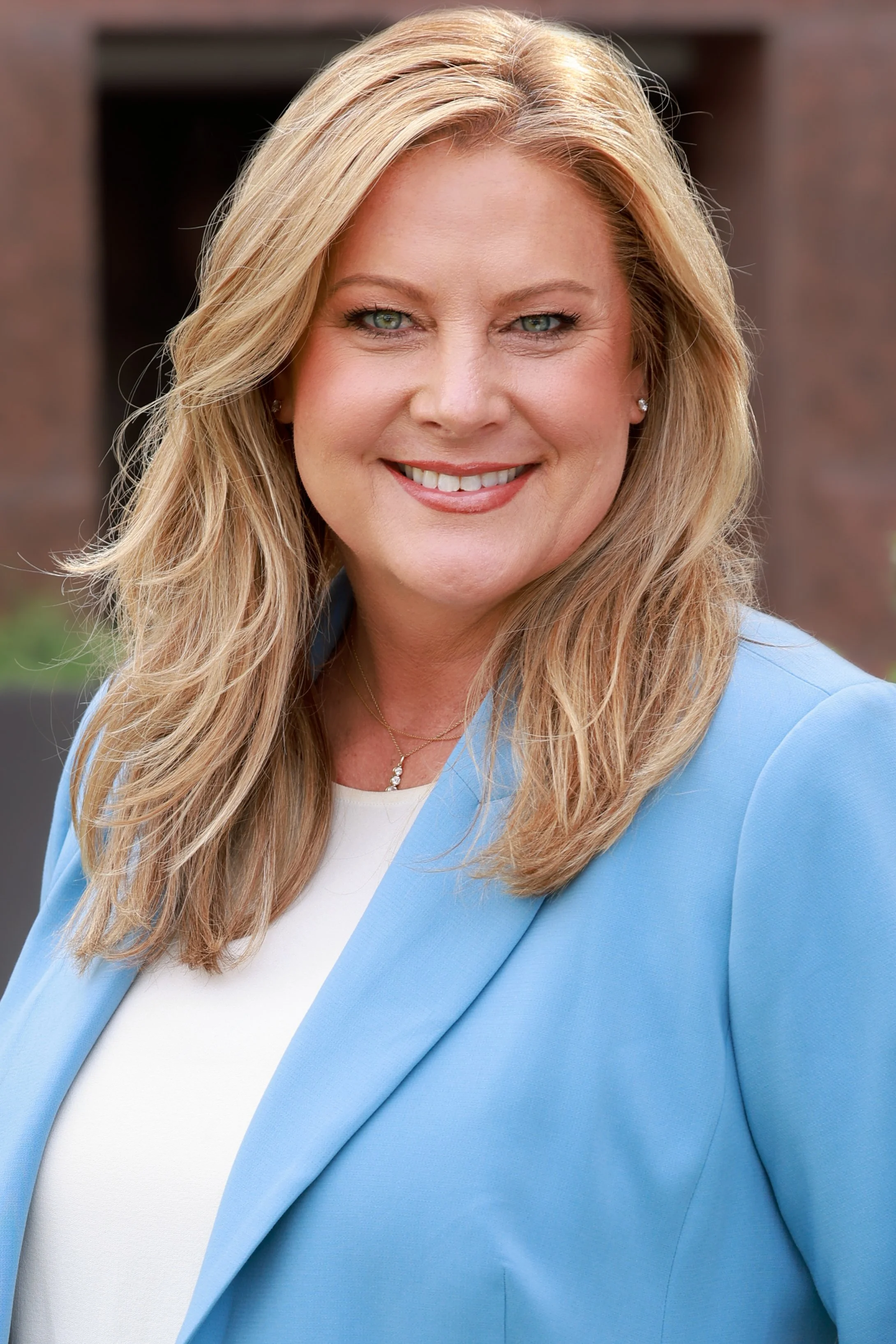 A smiling woman with blonde hair and blue eyes wearing a light blue blazer and a white top, standing outdoors in front of a brick building.