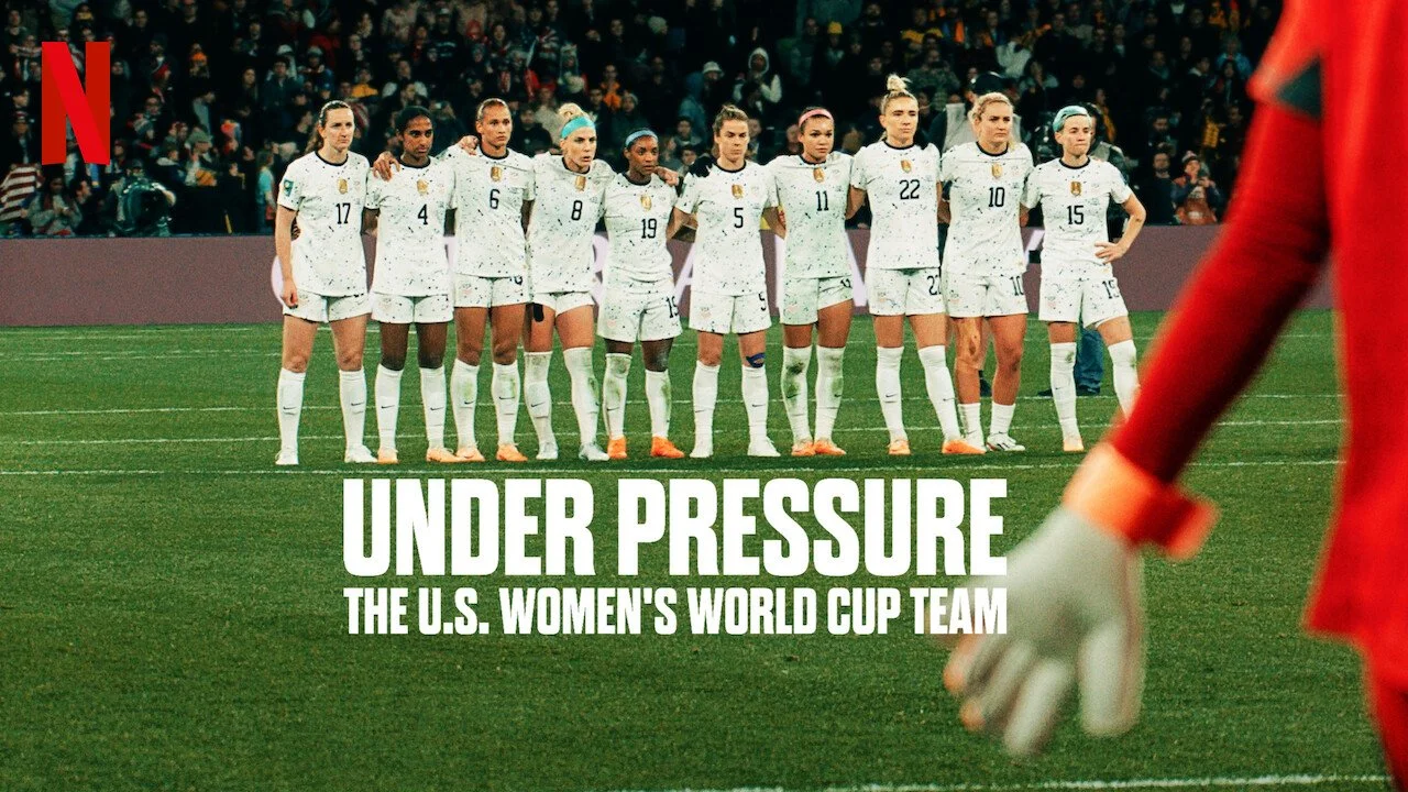 U.S. women's soccer team standing in a line on the field, wearing white uniforms. A goalkeeper in red is partially visible on the right. Crowd in the background. Text on image: 'Under Pressure, The U.S. Women's World Cup Team'.