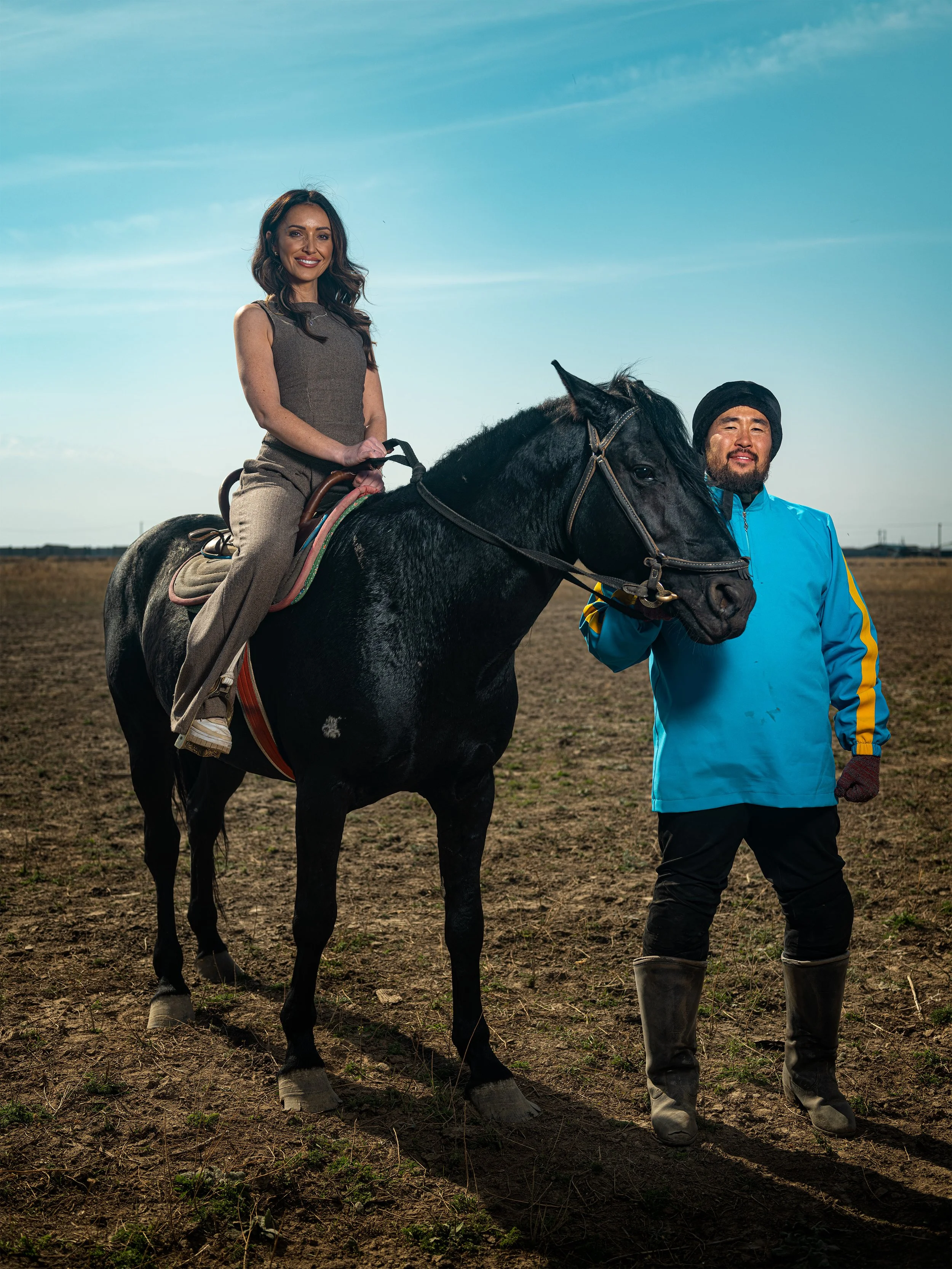 A woman riding a black horse outdoors on a clear day, accompanied by a man holding the horse's bridle. Lexus.
