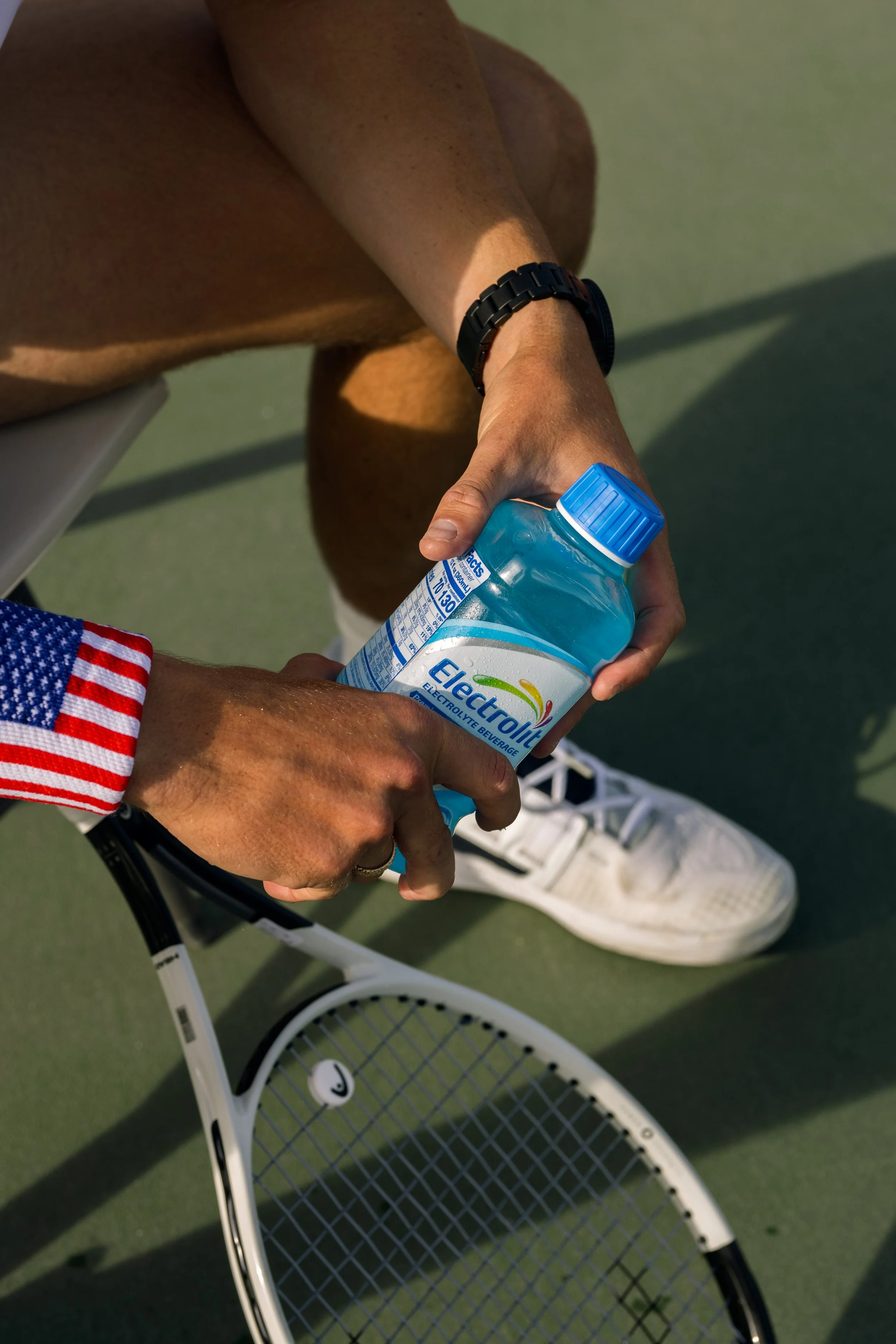 Person on a tennis court holding a bottle of electrolyte beverage and a tennis racket, wearing athletic shoes and a black watch.