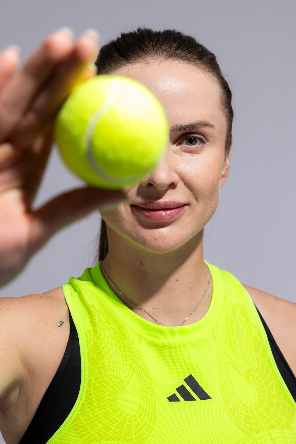 A woman holding a yellow tennis ball close to the camera, with her face in the background. She is wearing a bright yellow athletic top and has brown hair tied back. MacPaw