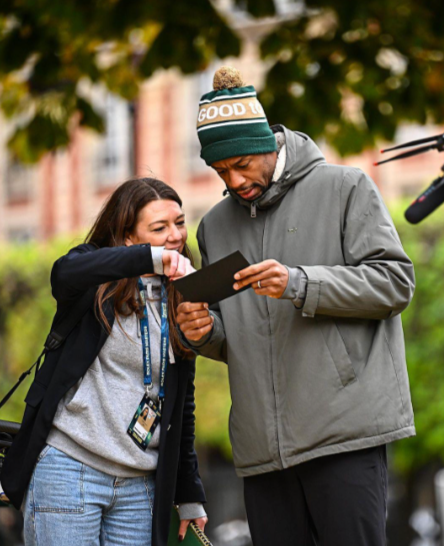 Two people, a woman and a man, looking at a smartphone together outdoors, with trees and buildings in the background.