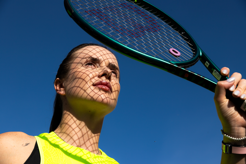 Close-up of a female tennis player holding a tennis racket with a shadow grid cast on her face, against a clear blue sky. McPaw