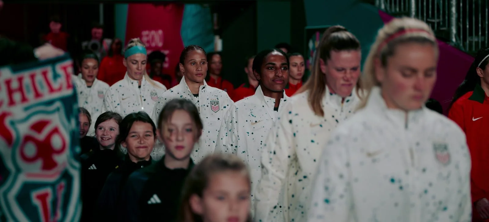 A group of female athletes walking in a line, wearing white jackets with USA patches, with children and other athletes in the background.