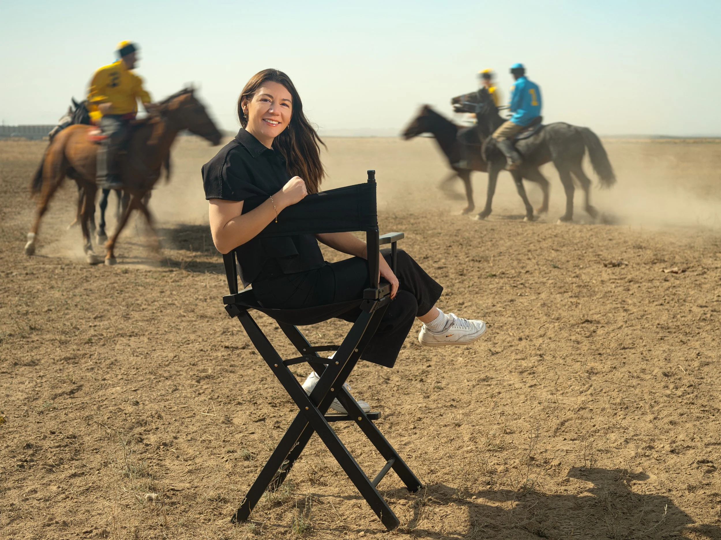 Angela Evans sitting on a director's chair in a dirt field, smiling at the camera, with a horse race happening behind her in a dusty, open landscape.