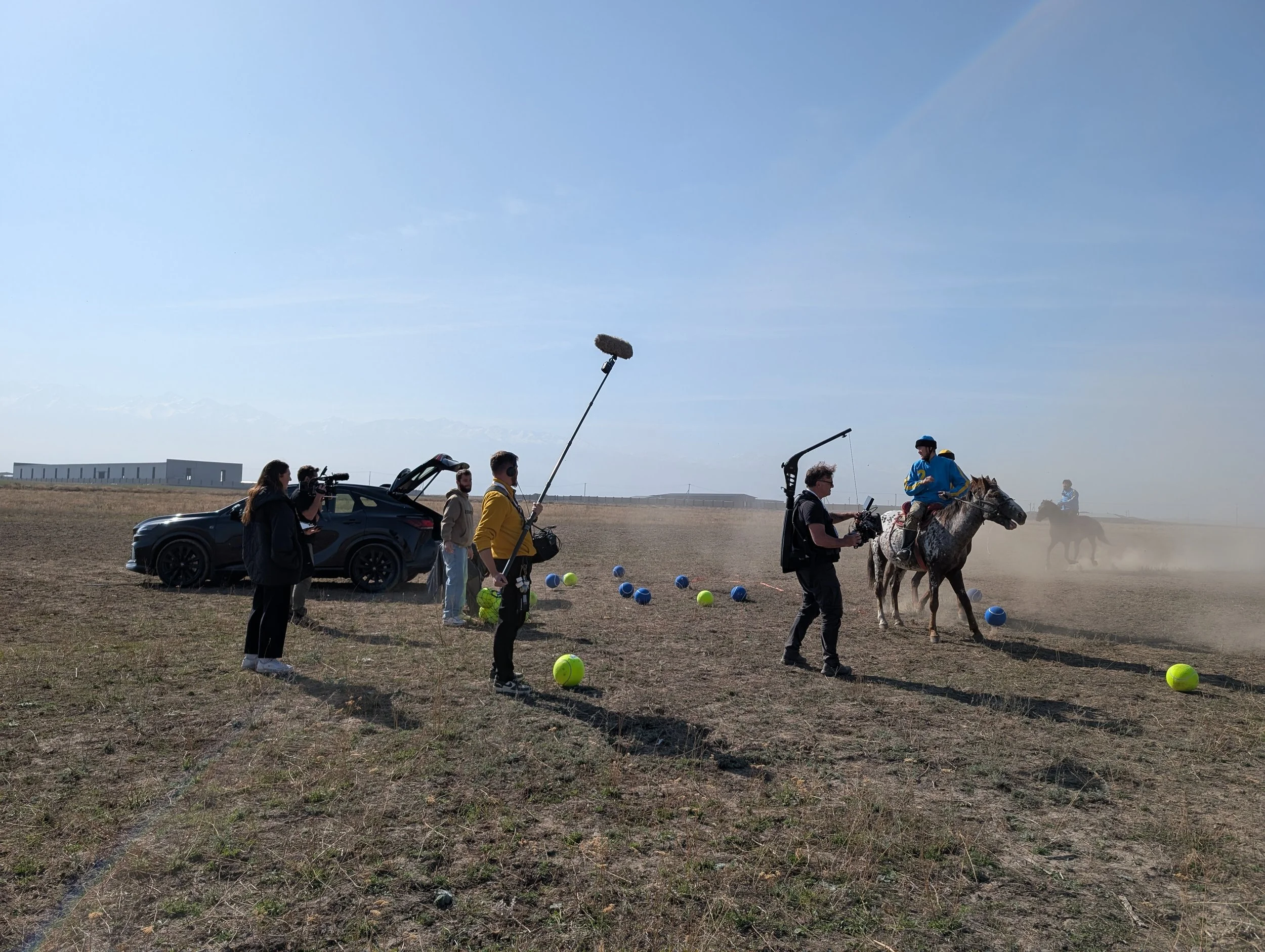 People filming a person riding a horse on a field with tennis balls scattered around, some people on horses in the background, and a car parked nearby. Lexus.