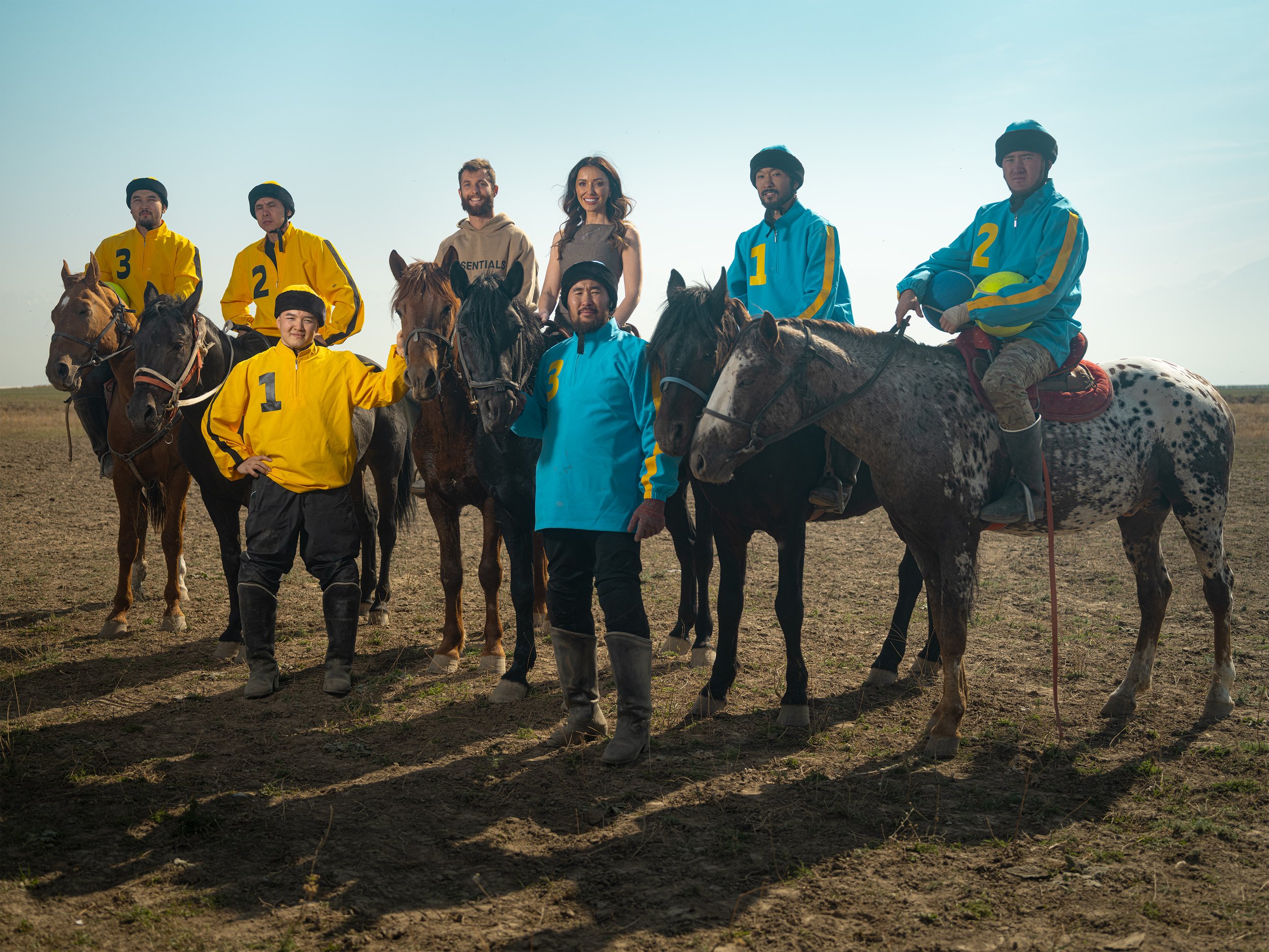 Group of seven people and eight horses on an open field, some riders wearing numbered jackets, with clear sky in the background. Lexus 2025.