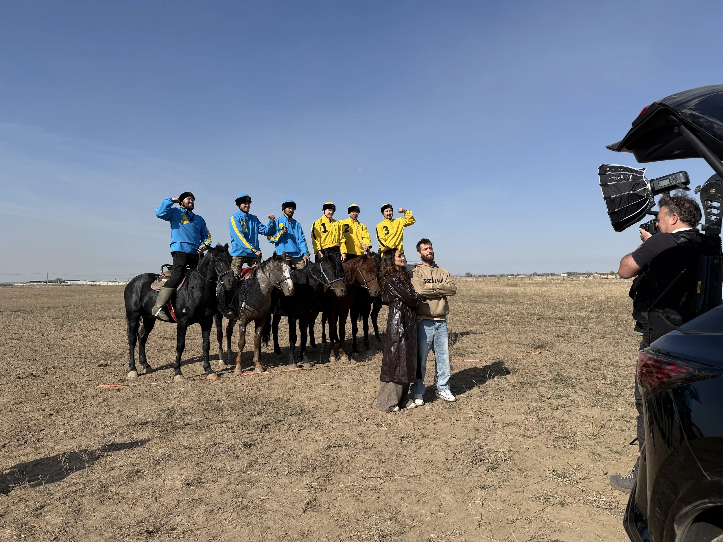 Group of six horseback riders, three in blue and three in yellow jerseys, on a flat open field during a photo shoot. Two women and a man stand in front of the horses, and a photographer is taking pictures from a vehicle on the right side. Lexus.