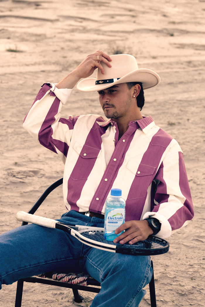 A man sitting outdoors on a desert-like terrain, wearing a oversized pink and white striped shirt, a white cowboy hat, and holding a tennis racquet rest on his lap with a water bottle labeled 'Electrolit' on the racquet. The man has dark hair, a small goatee, an earring, and appears to be gazing thoughtfully into the distance.