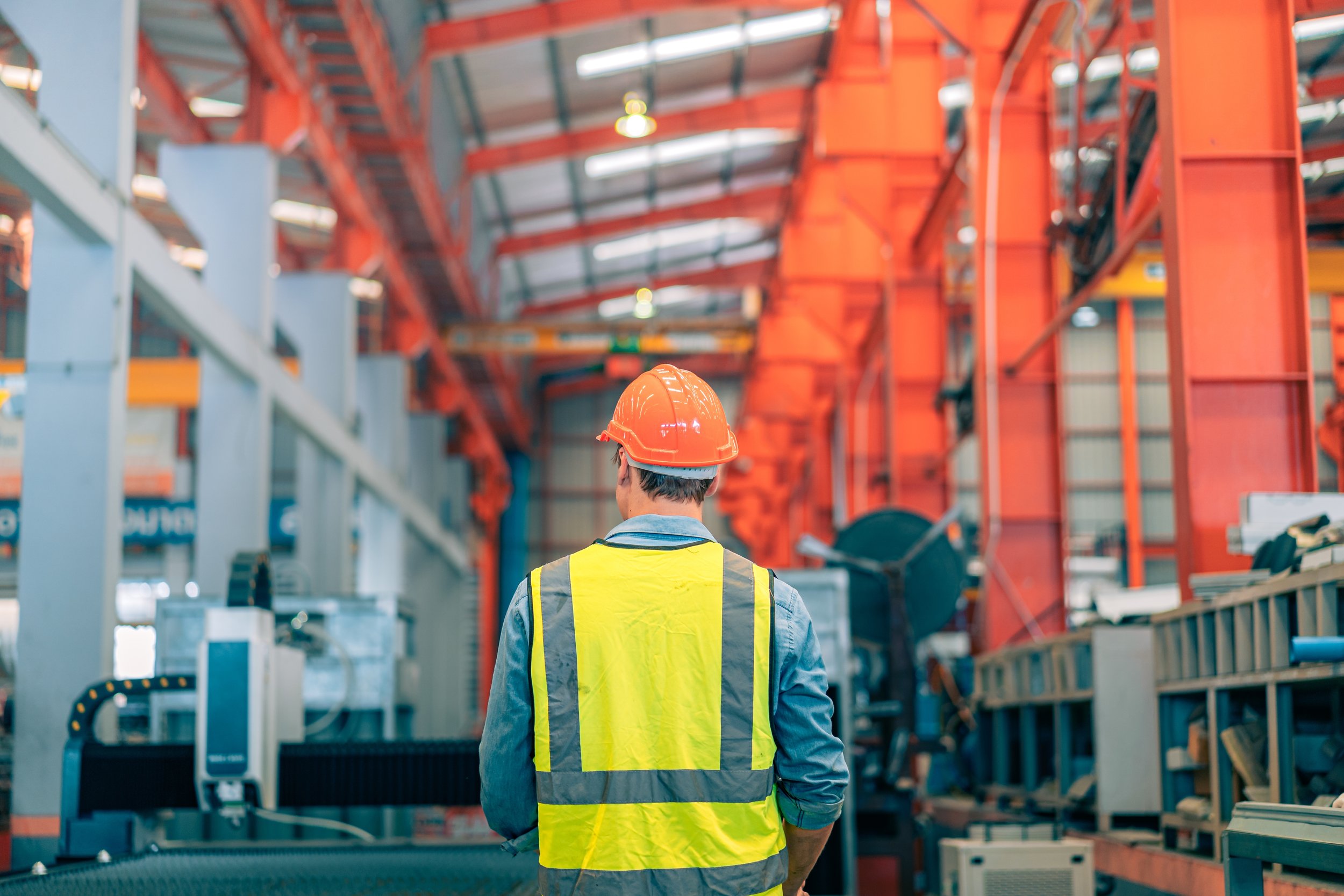 A person wearing a yellow safety vest and an orange hard hat stands in a warehouse or industrial setting, facing away from the camera. The background shows orange and grey shelving units with various equipment and materials.