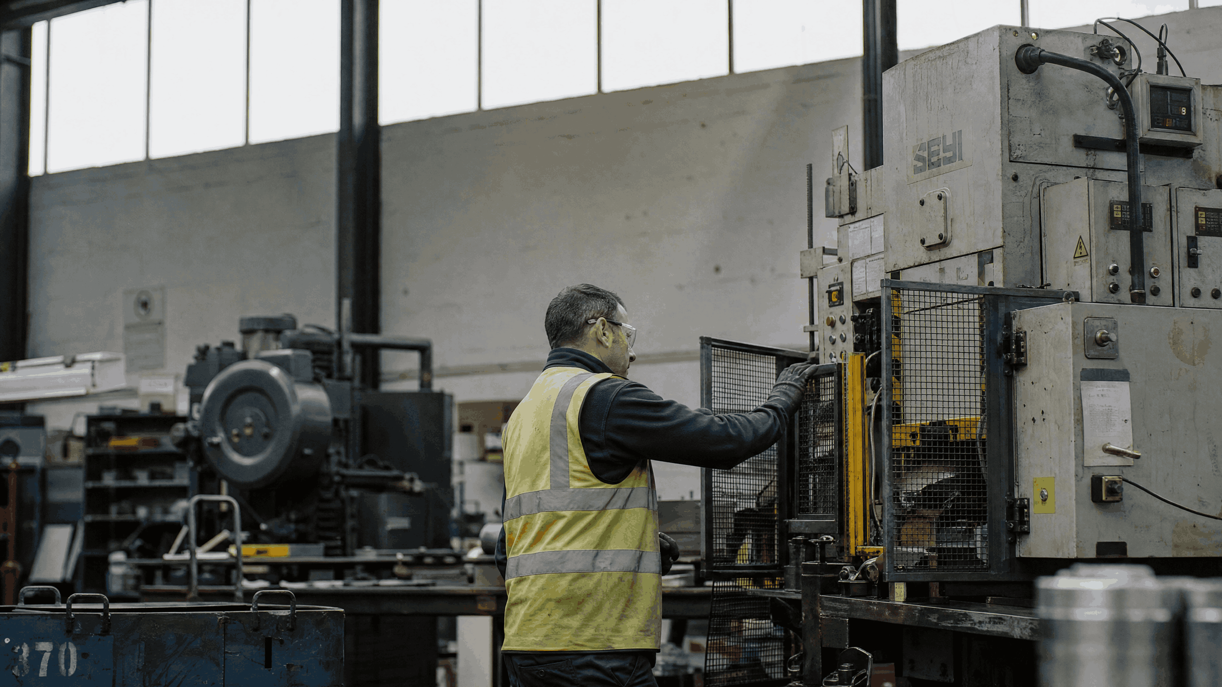 A worker wearing safety glasses and a yellow safety vest operating industrial machinery in a factory setting with large windows.