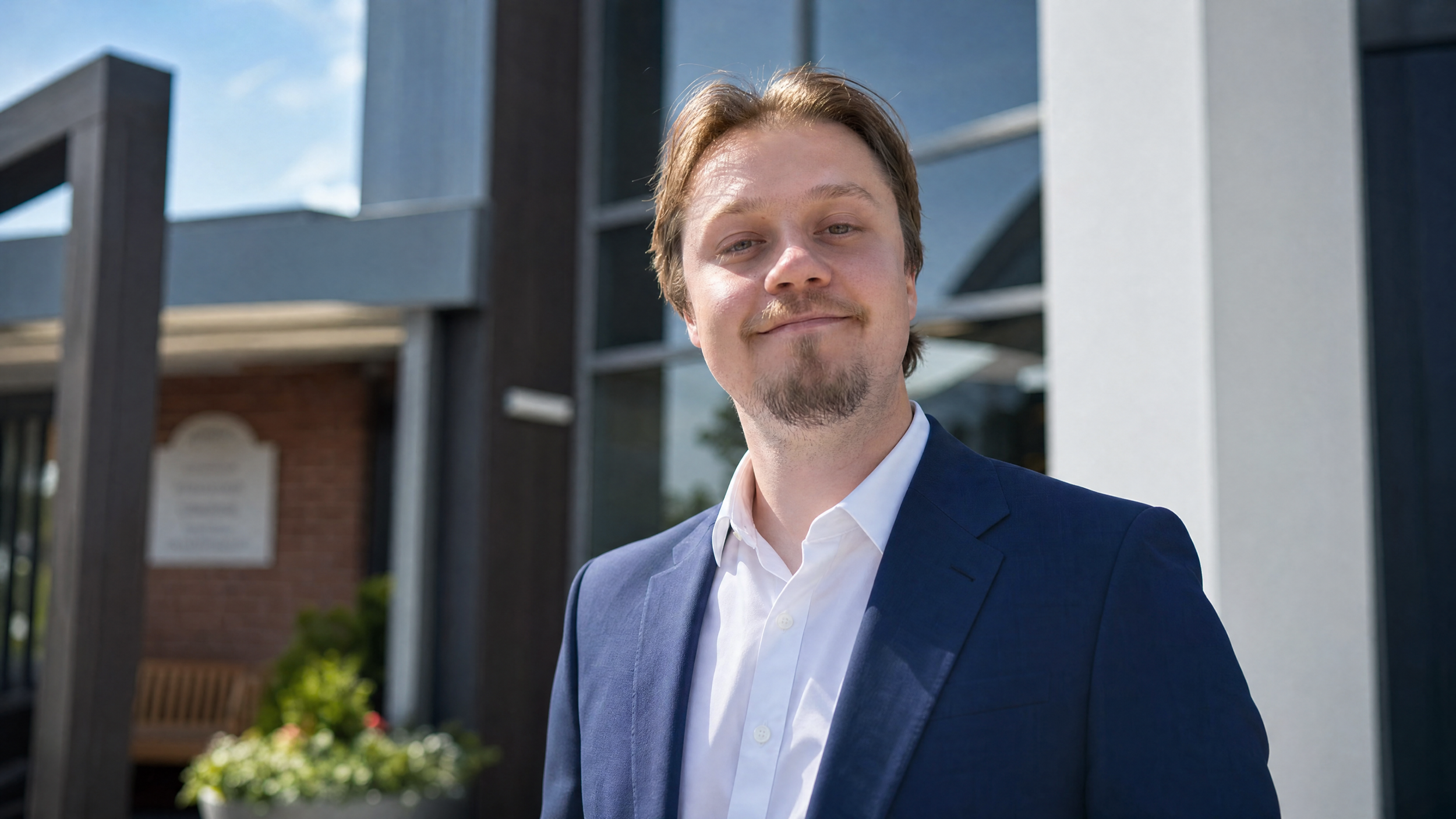 A young man with light brown hair, a slight mustache, and goatee wearing a navy suit and white shirt, standing outdoors in front of modern buildings.