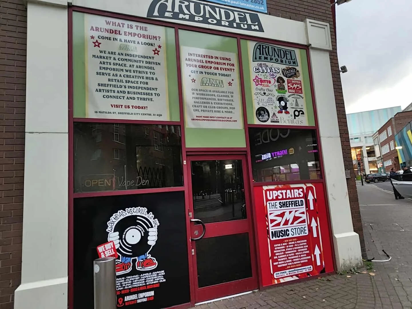 Storefront of Arundel Emporium with multiple signs, including a large red and white sign indicating 'Upstairs The Sheffield Music Store,' and informational posters about the store's community space, events, and contact details.
