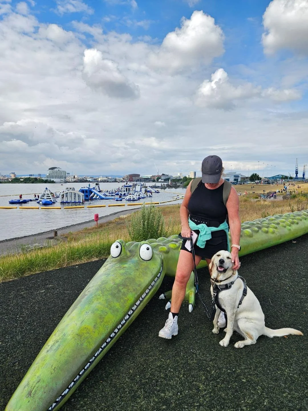 A woman in black athletic clothing and a gray cap standing on a paved path next to a sitting Labrador retriever dog, with a green crocodile-shaped bench and a waterfront with inflatable water attractions and a city skyline in the background.