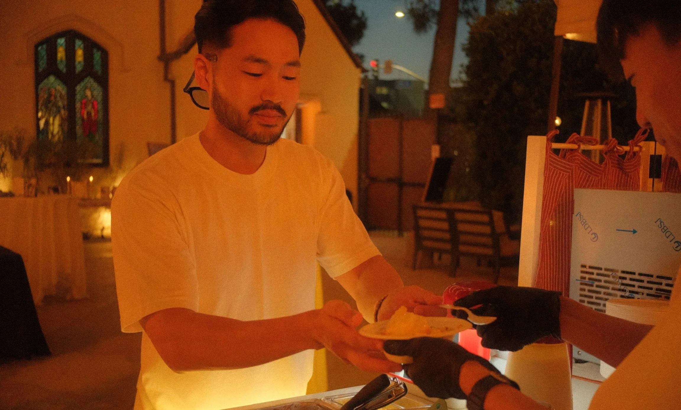 A man in a white t-shirt is at a counter, receiving a bowl of Korean shaved ice from another person. The background shows a church with stained glass windows and lit candles, suggesting an evening wedding setting.