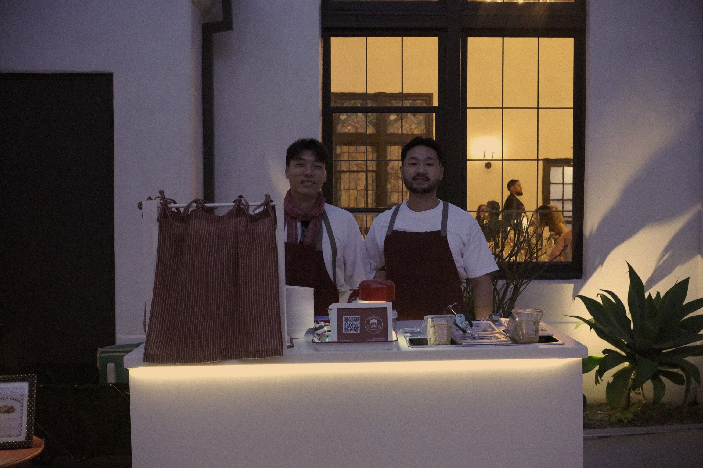 Two men standing behind a white mobile shaved ice bar at a wedding venue during evening, with house-like windows and warm lighting visible behind them.