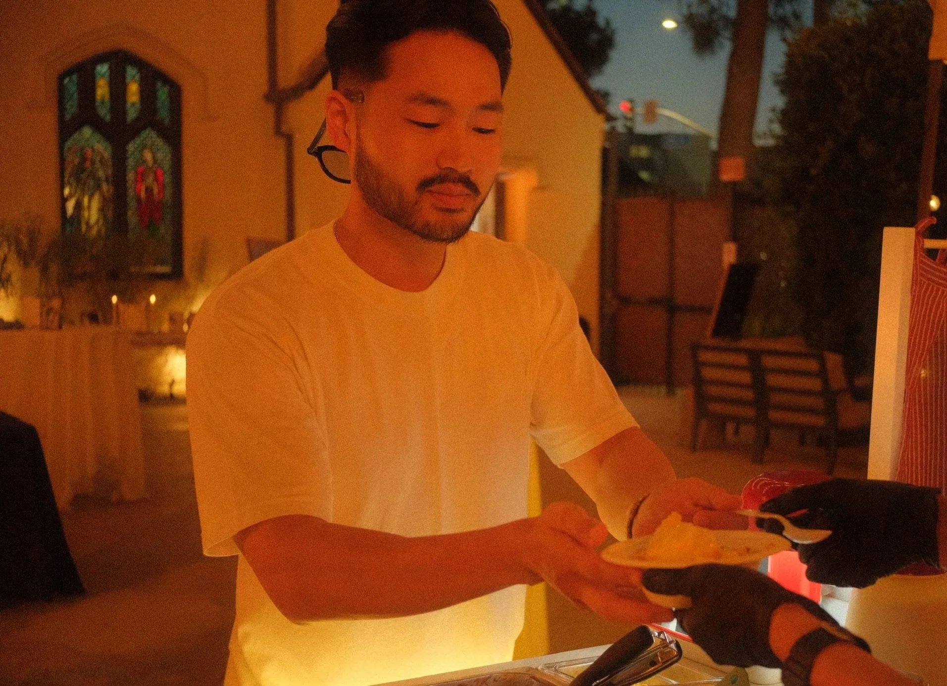 A man in a white t-shirt is at a counter, receiving a bowl of Korean shaved ice from another person. The background shows a church with stained glass windows and lit candles, suggesting an evening wedding setting.