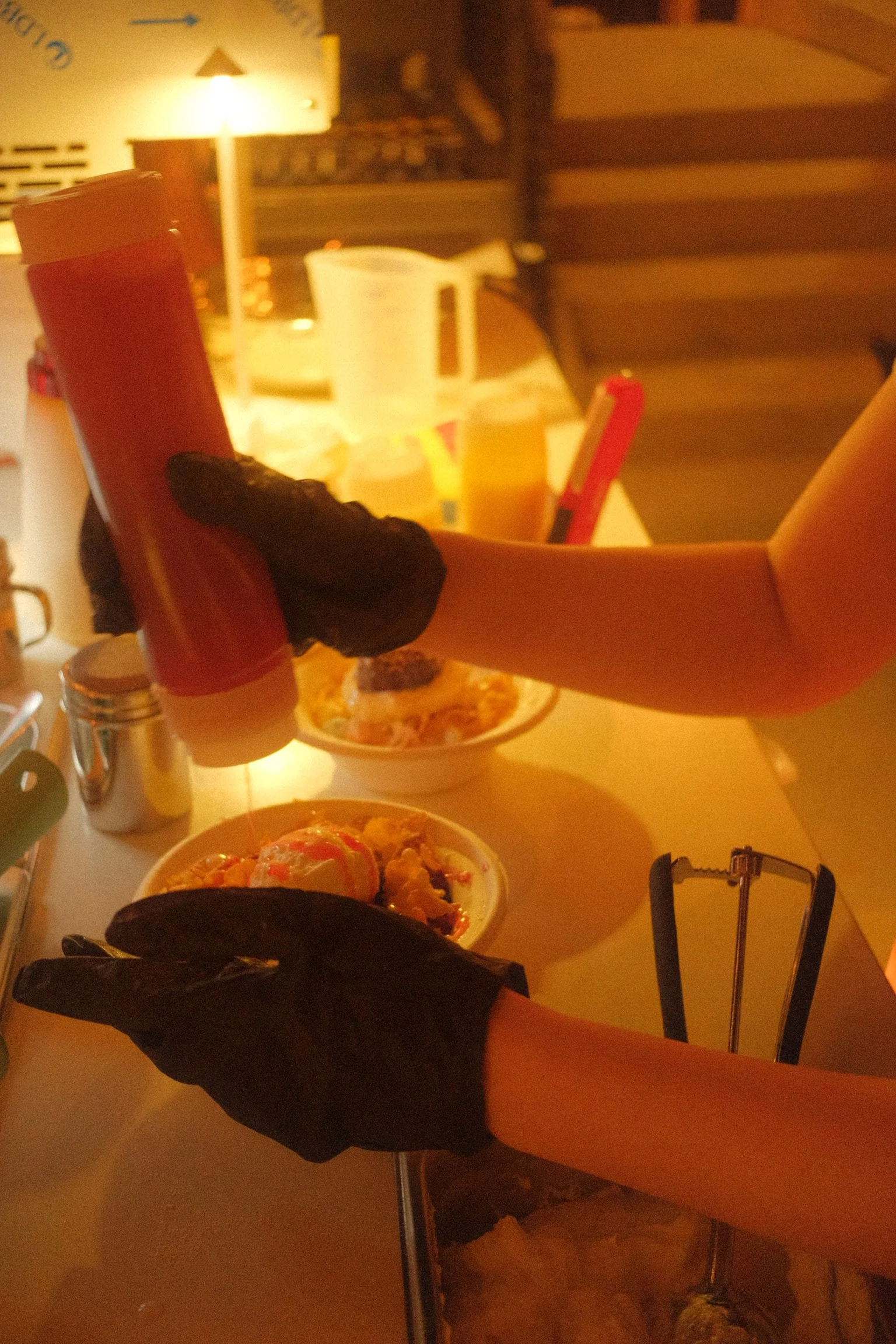 Two people are using black gloves to hold a burger with a bun, leaf lettuce, tomato, cheese, and possibly a patty. In the background, there are food dishes, drinks, and condiments on a table, with a warm, cozy atmosphere.