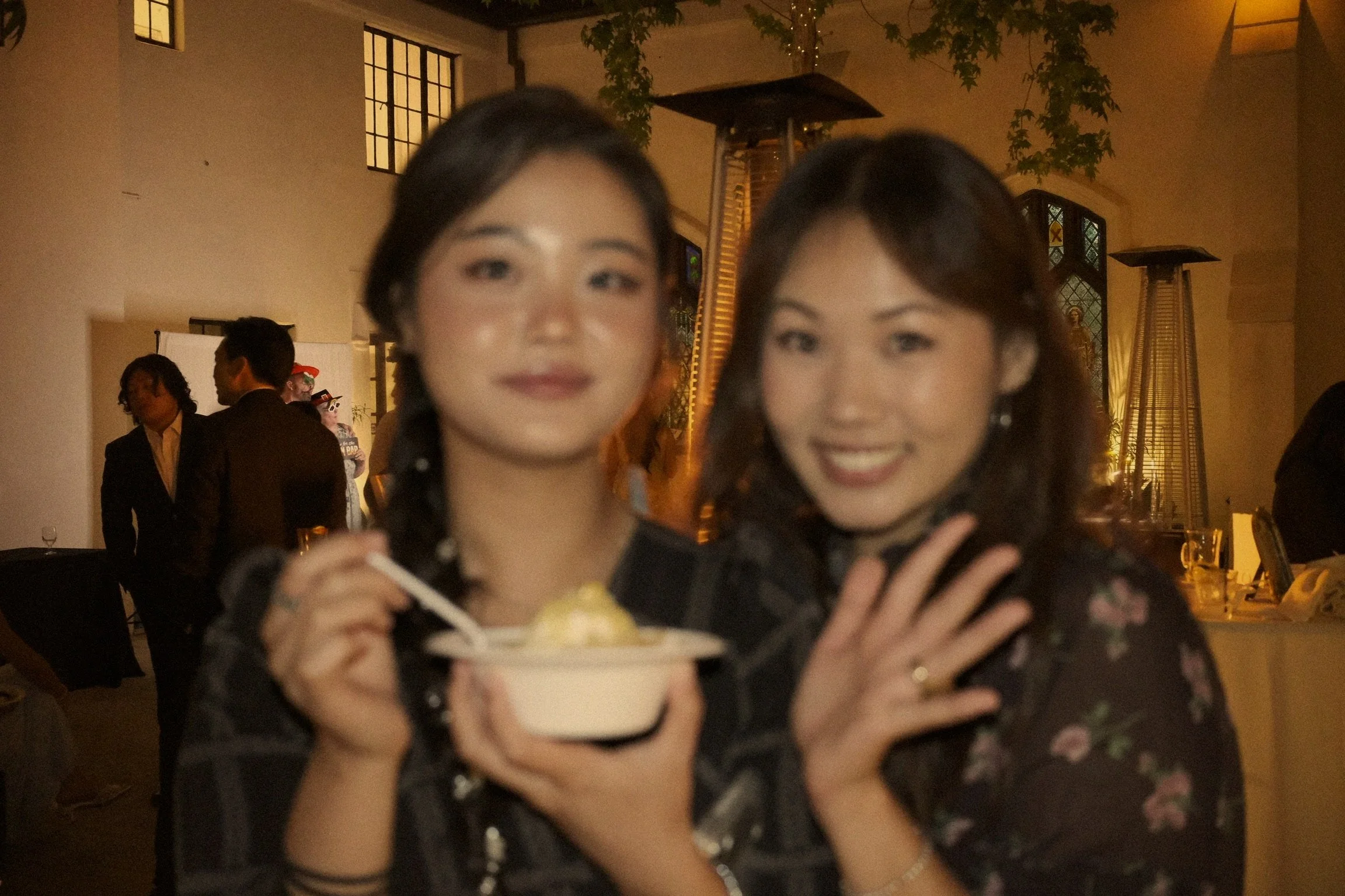 Two women with dark hair and floral-patterned clothing smiling at a celebration, one holding a bowl of shaved ice dessert, with a group of people in the background in an indoor venue with warm lighting and windows.