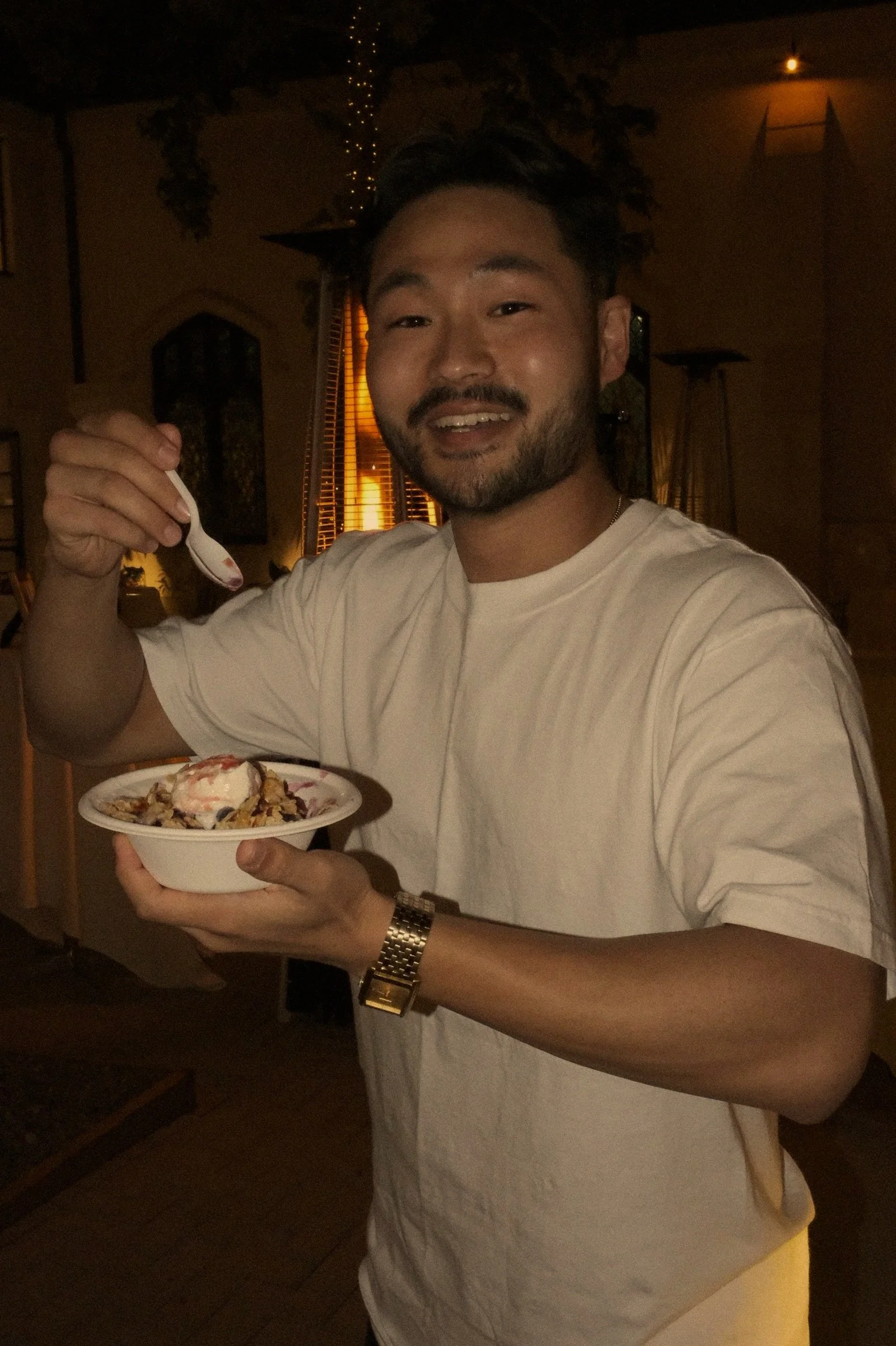 A young man with dark hair, a beard, and a white shirt is smiling and holding a bowl of Korean shaved ice with toppings, in a dimly lit indoor setting.