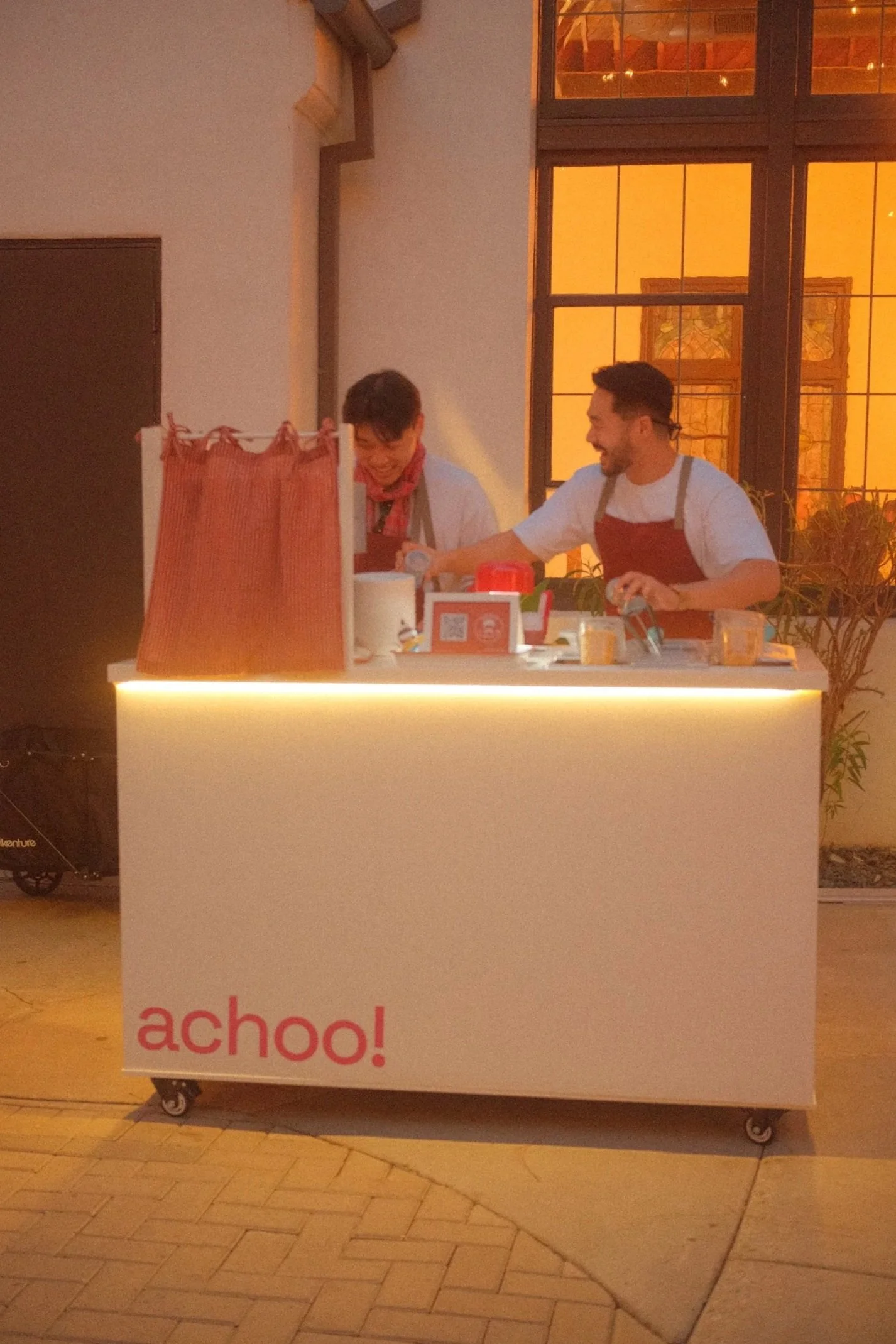 Two people smiling and interacting behind a counter with a privacy screen, inside a warmly lit space with large windows.