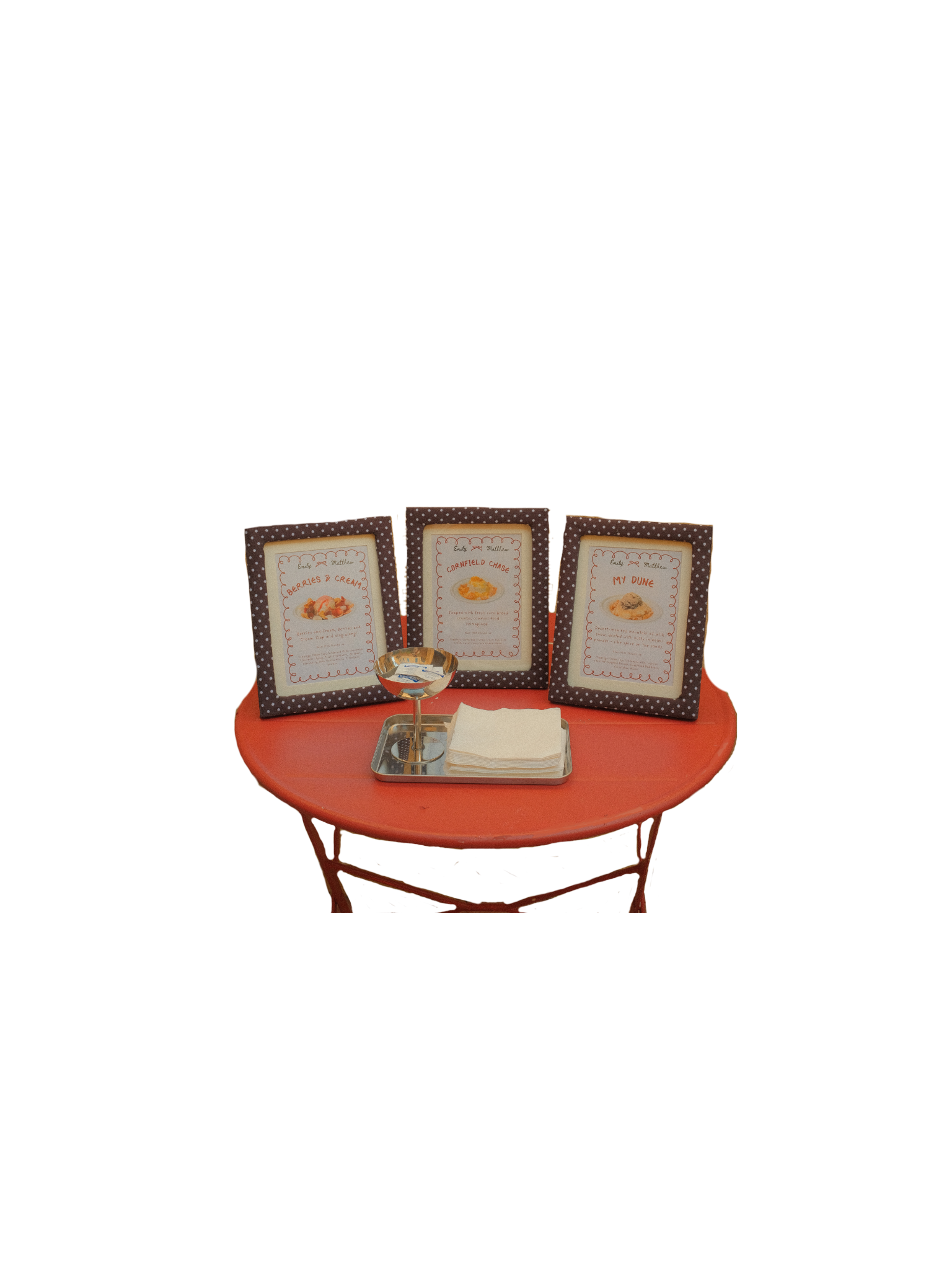 Display of three framed photographs of desserts, silver bowl, and white napkins on a red round table