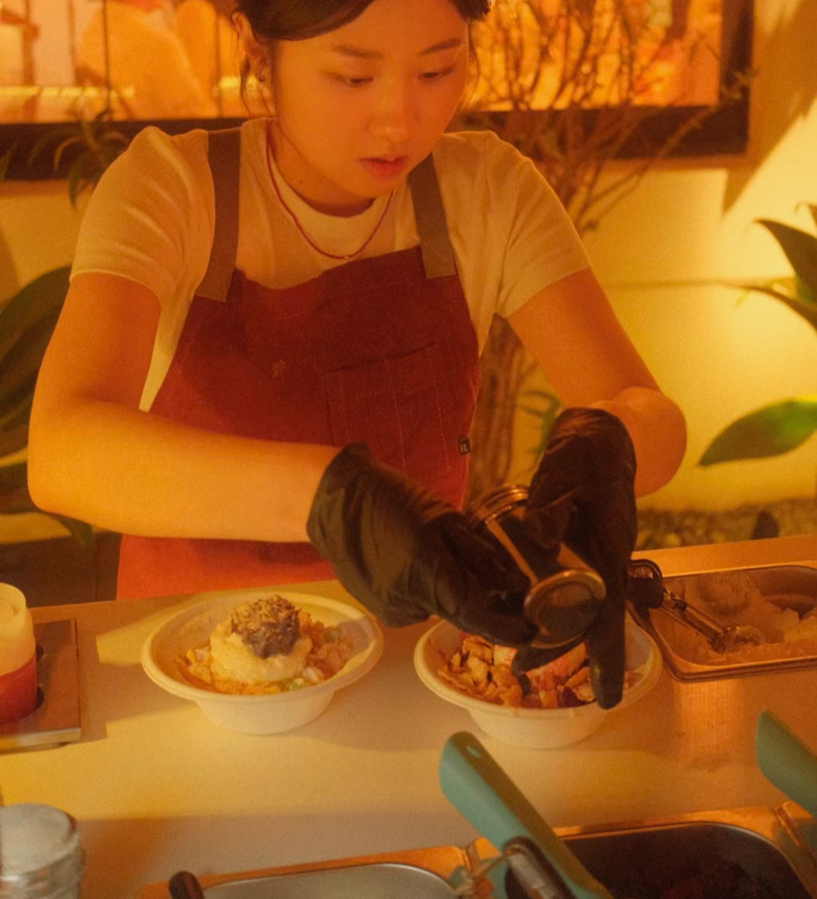 A young woman wearing a red apron and black gloves is preparing food in a restaurant setting, with plates of food in front of her.
