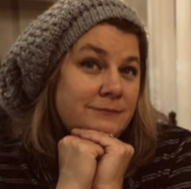 Amanda Pfennig Second Helping Avenue A Films Close-up of a woman with shoulder-length brown hair wearing a gray knit beanie and a striped shirt. She is resting her chin on her hands and smiling softly.