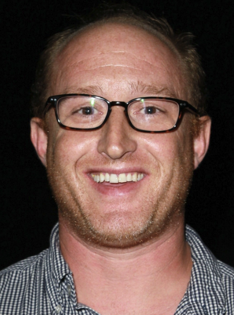 Hunter Bodine Avenue A Films Second Helping A smiling man with glasses, short light brown hair, and a beard, wearing a checkered shirt, against a black background.