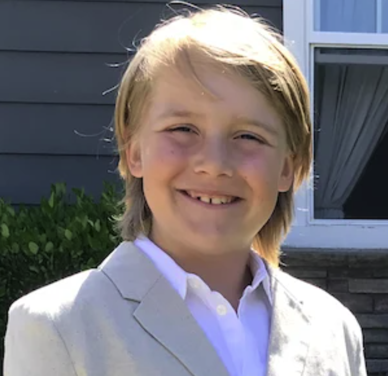 Smiling young boy with blonde hair in a white suit and shirt, standing outside near a house with a window and shrubbery.