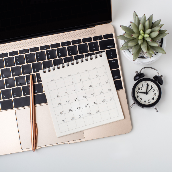 A workspace featuring a closed laptop, a rose gold pen, a blank desk calendar, a small potted succulent plant, and an analog alarm clock showing 11:51.