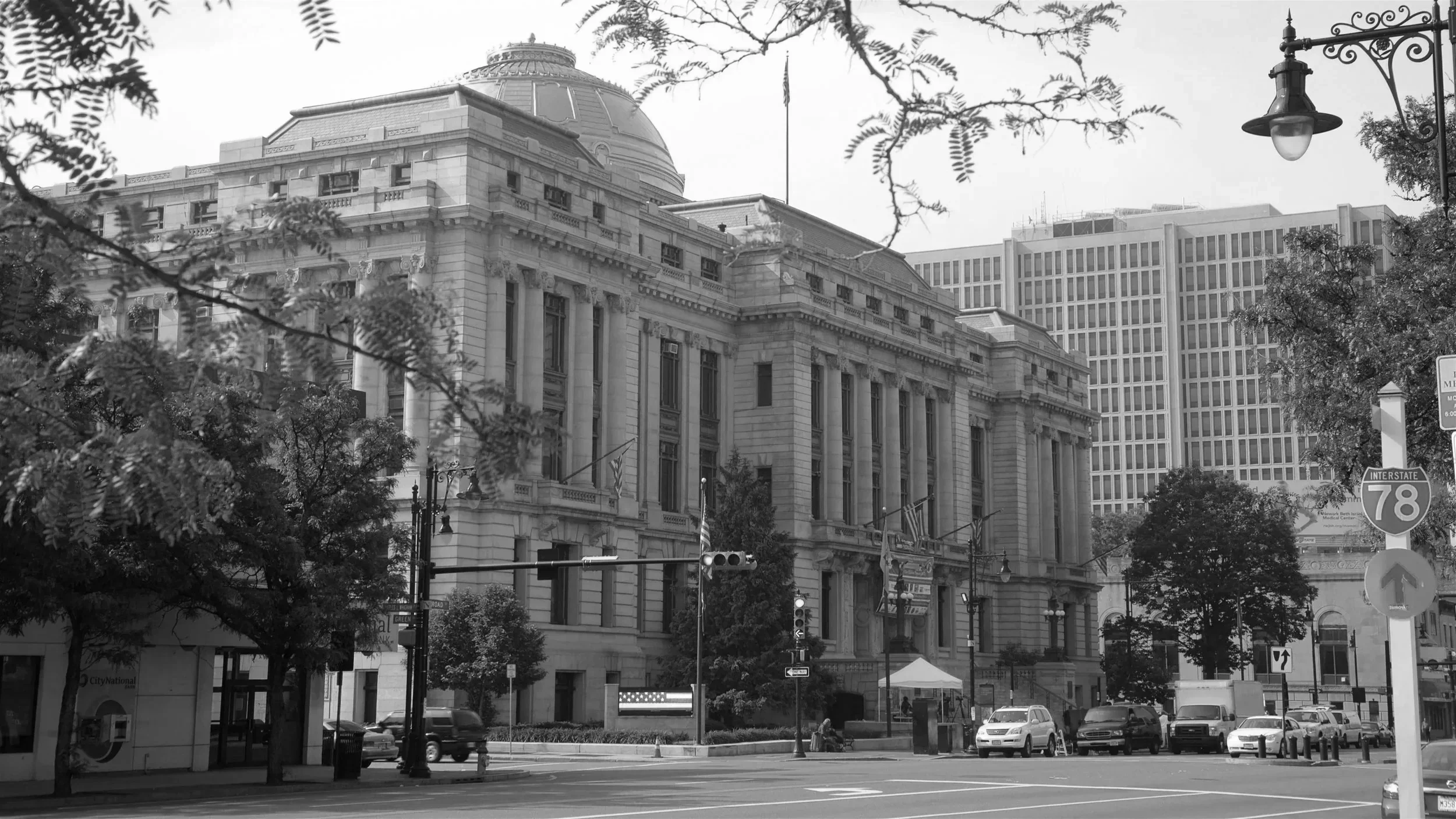 Black and white photo of a historic government building with classical architecture, large columns, and a dome, situated on a city street with trees, cars, street signs, and a lamppost.