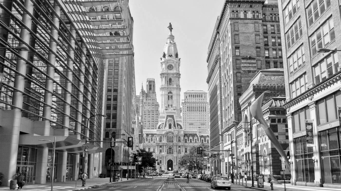 Black and white photo of a city street with tall buildings, leading to a historic clock tower with a statue on top.