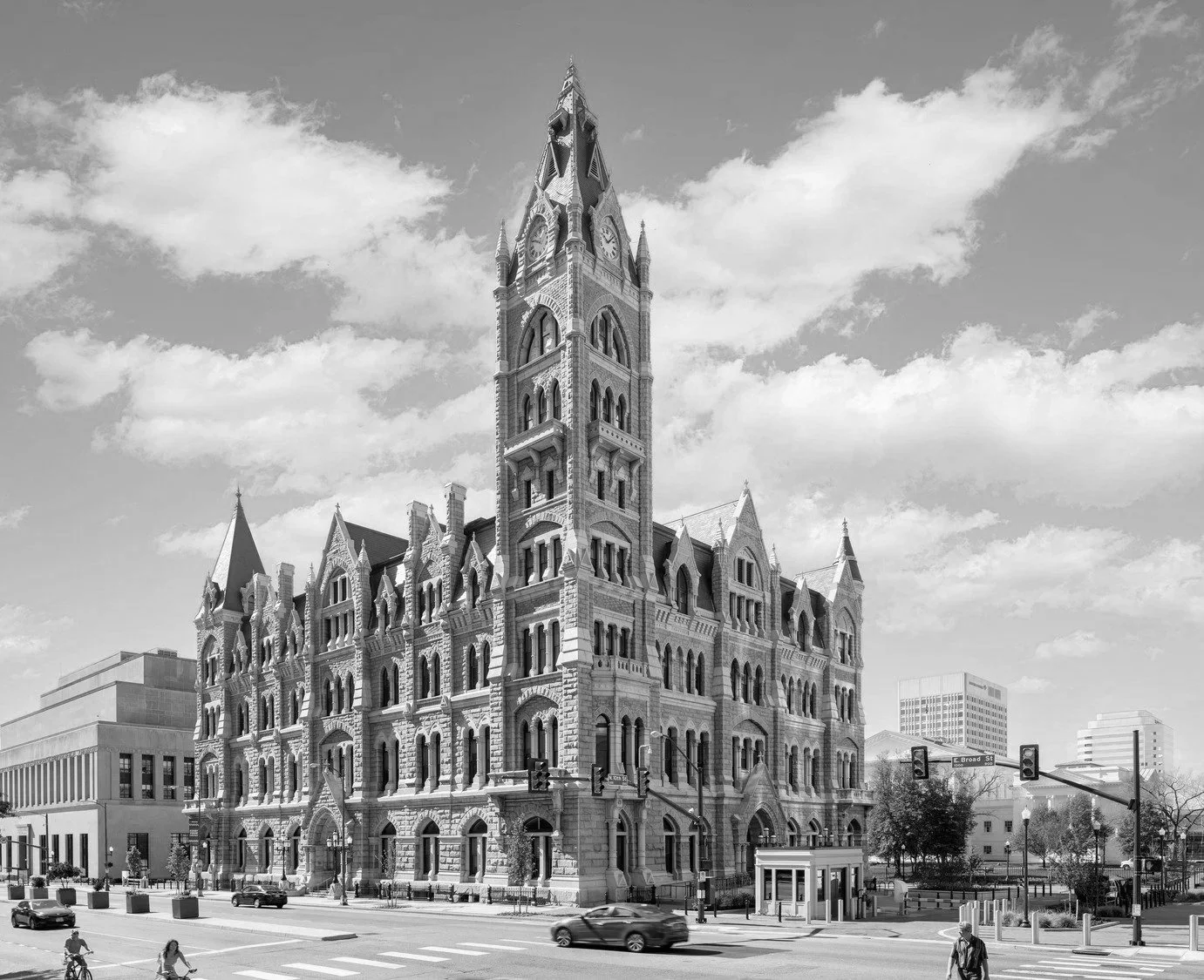 A black and white photo of a historic stone building with a tall clock tower, located at an intersection with traffic lights and cars passing by.