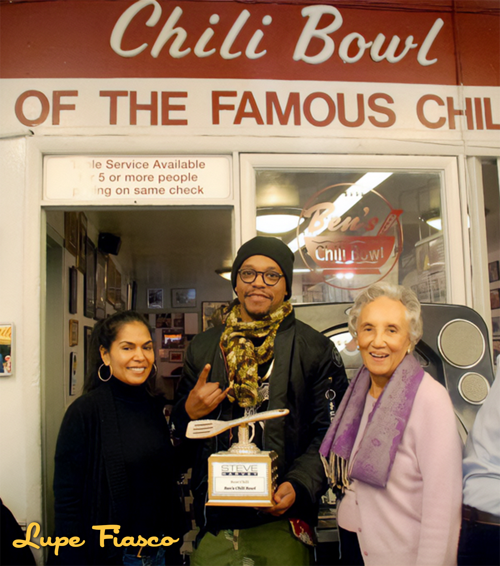 Three people standing in front of a restaurant called Chilli Bowl. The person in the middle holds a trophy and makes a rock sign with one hand. The woman on the left is smiling, and the older woman on the right is also smiling. The background shows t