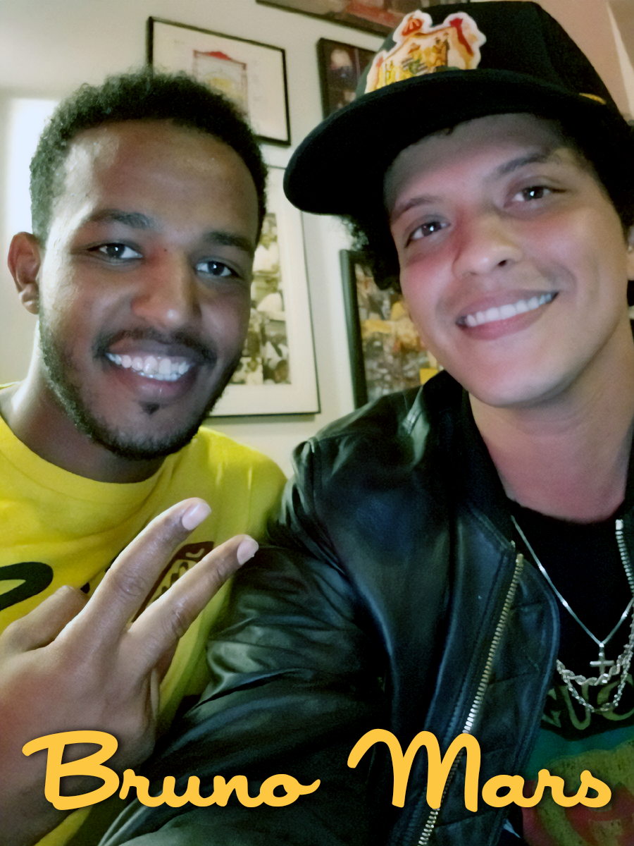 Two young men smiling, one wearing a black leather jacket and a hat with a colorful patch, the other in a yellow shirt showing a peace sign, indoors with framed pictures on the wall.