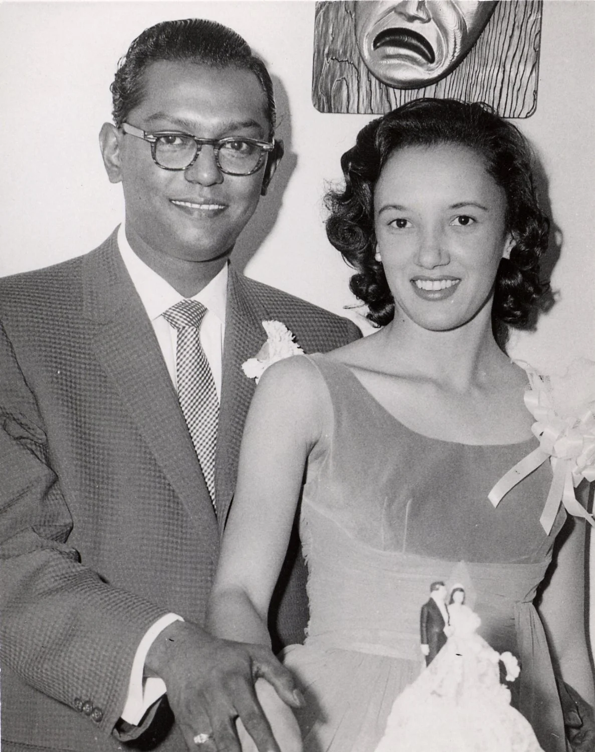 Black and white photo of a man and a woman smiling at a wedding, with a bride and groom miniature figurine on her dress, and an art piece with a crying face hanging on the wall behind them.