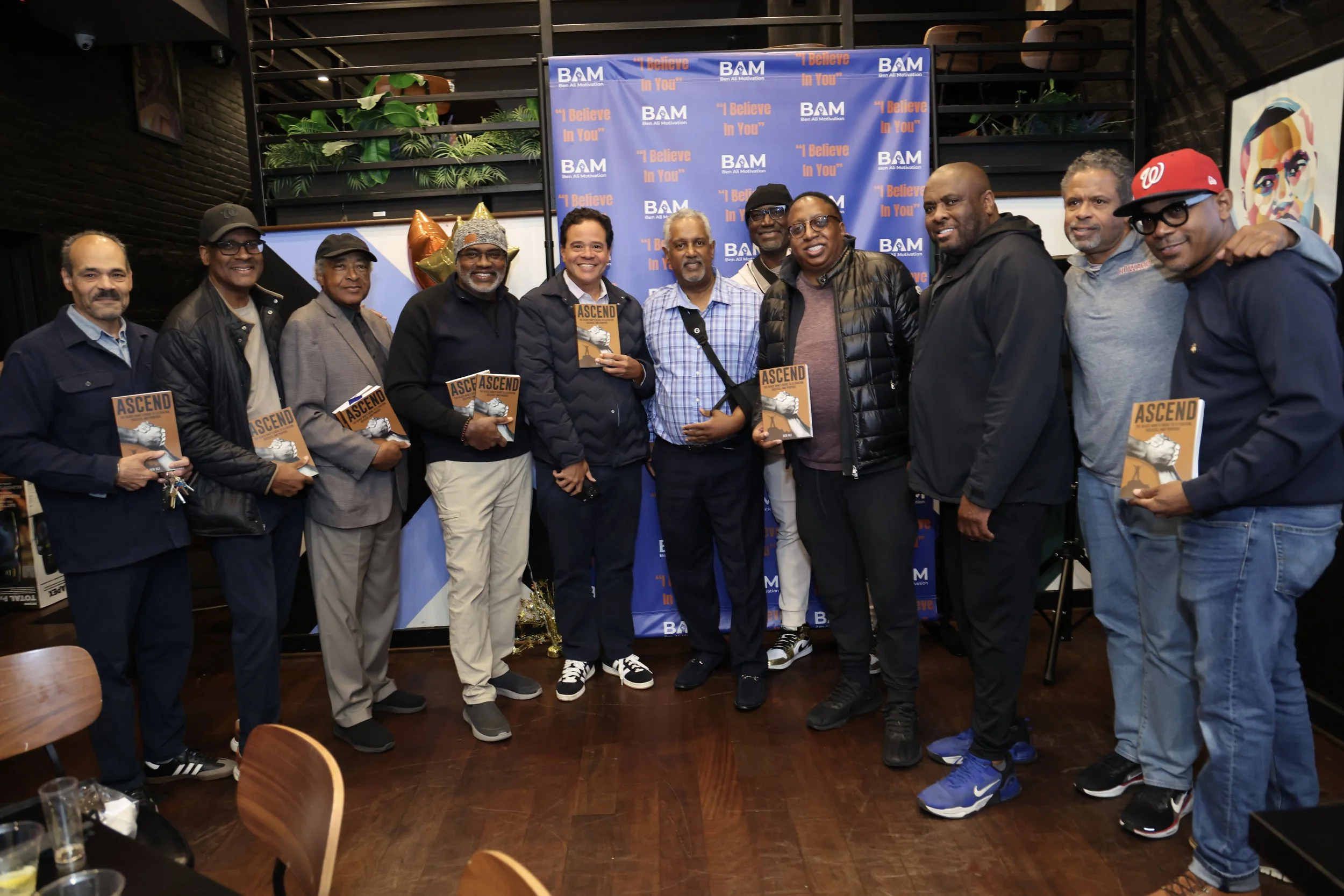 A group of people posing for a photo at a book launch event, holding copies of the book titled 'Ascend'. The background features a blue banner with the logo 'BAM' and the phrase 'I Believe In You'. The setting appears to be indoors with decorative elements and artwork on the walls.