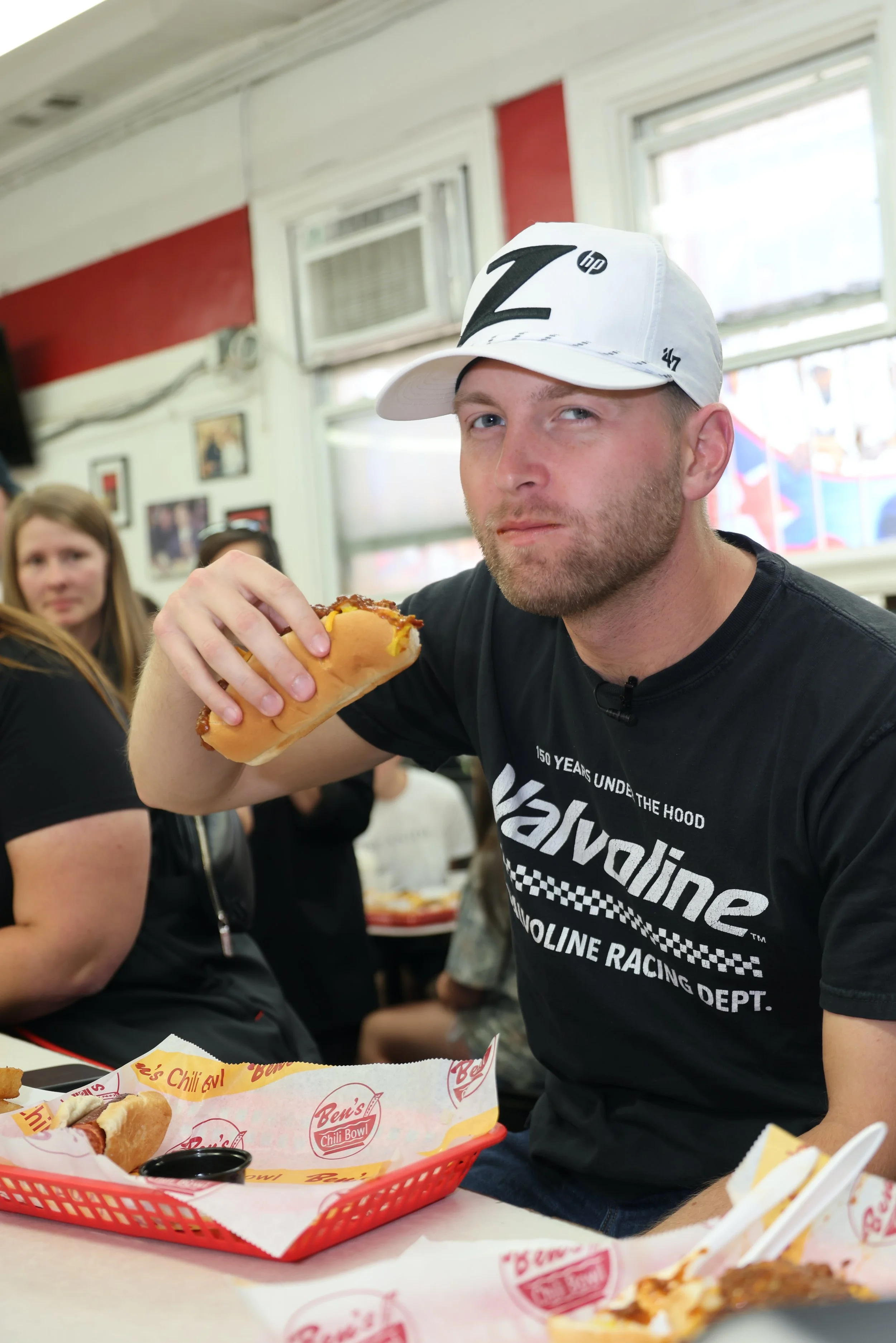 Young man with a beard wearing a white cap and a black T-shirt sitting inside a restaurant, holding a hot dog with chili. There are other people in the background and food on the table.