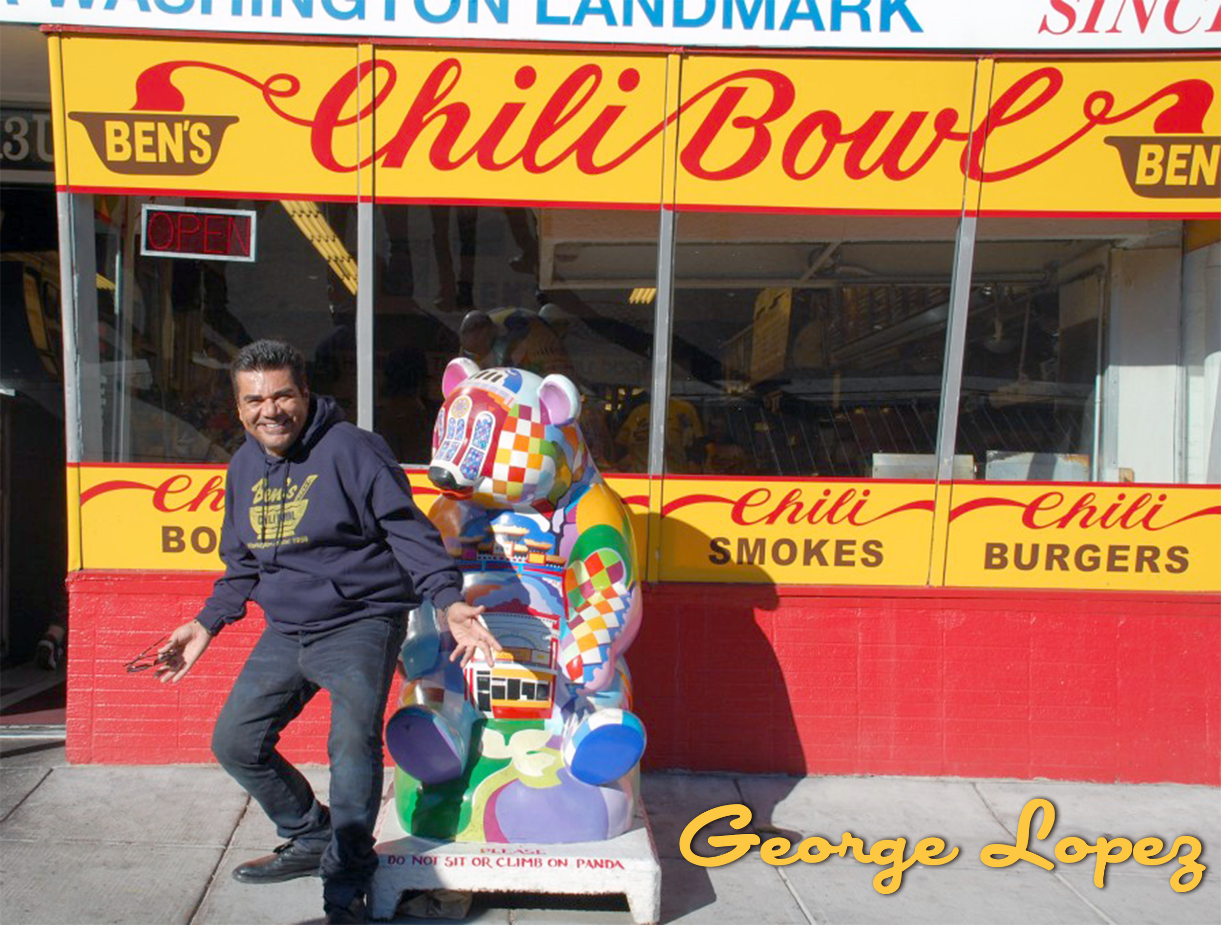 A man standing next to a colorful, decorated panda sculpture outside a restaurant named Ben's Chili Bowl. The restaurant's signage is yellow with red writing, advertising chili, smoked items, and burgers. The man is smiling, wearing a navy blue sweat