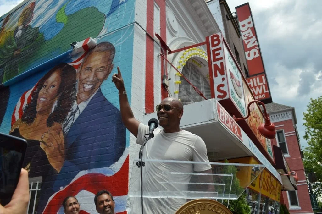 A man smiling and gesturing with his hand while speaking at a podium outdoors in front of a mural of two smiling people. The mural is painted on a brick building with signage for Ben's. The sky is cloudy.