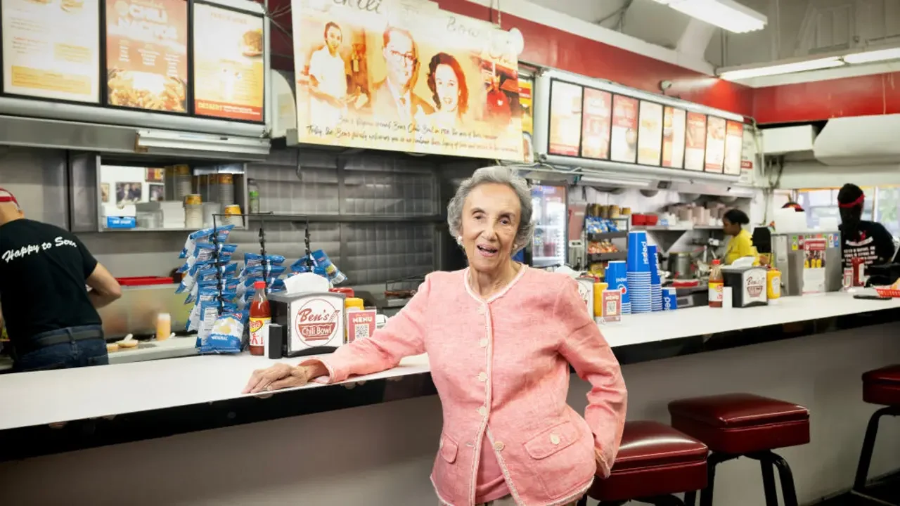 An elderly woman smiling inside a fast food restaurant, leaning on the counter with her right hand, with employees working behind the counter.