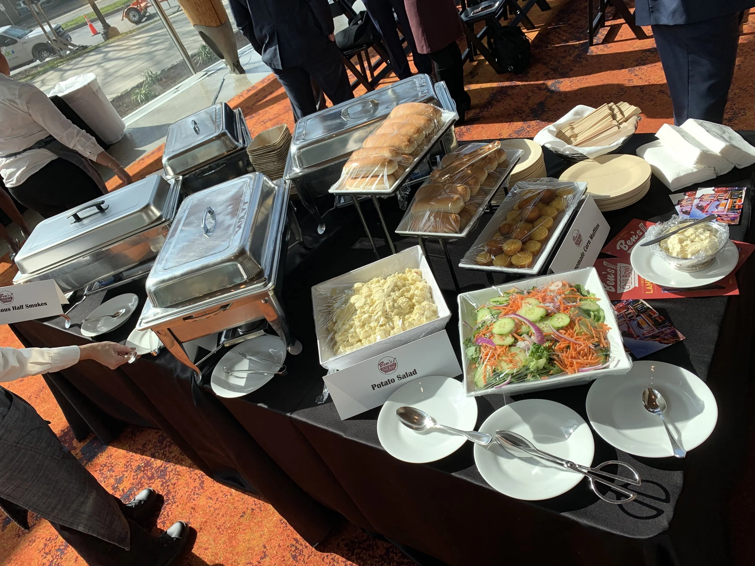 Buffet table with chafing dishes, bread rolls, potato salad, vegetable salad, and assorted sides at a catered event