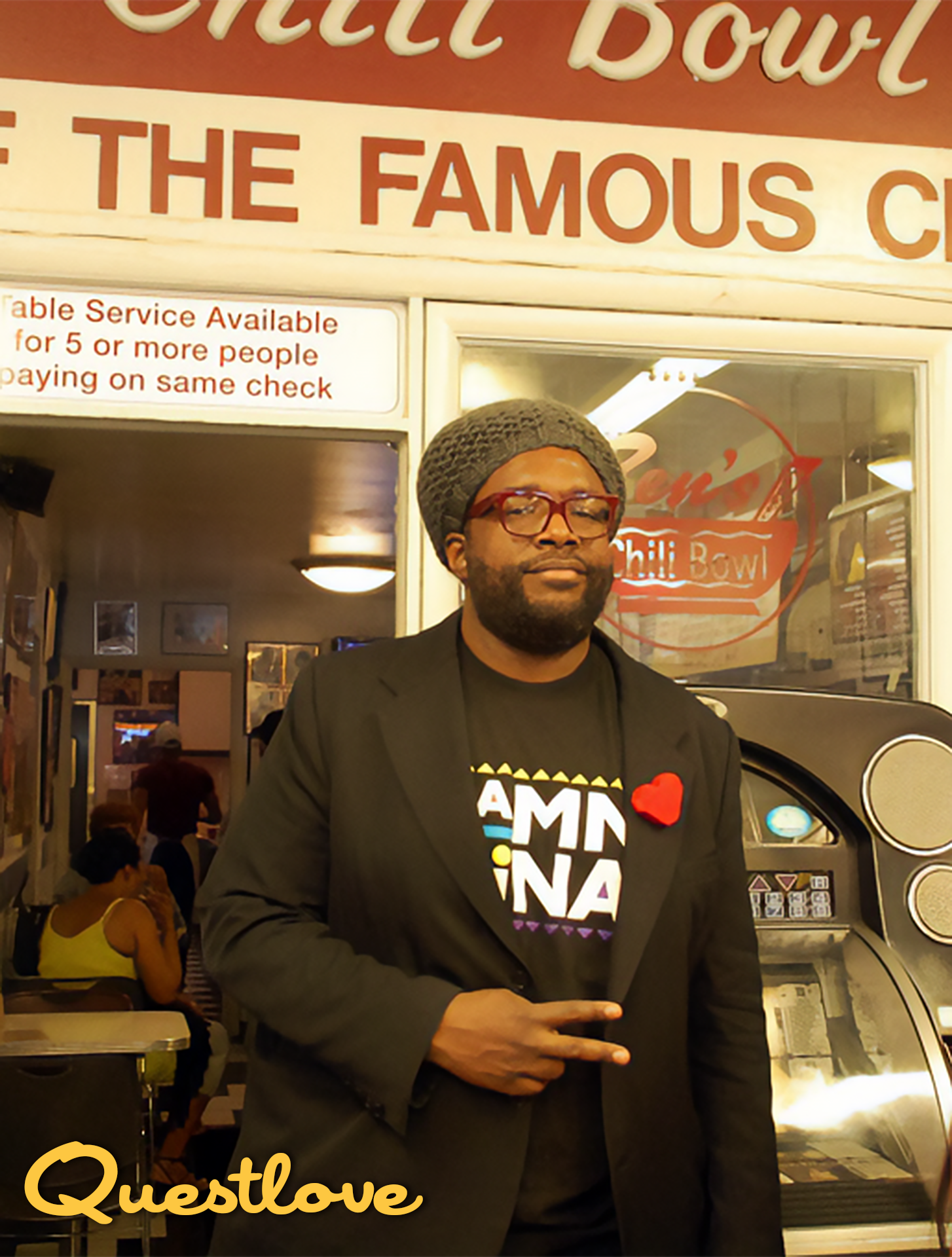 A man with glasses, a beard, and wearing a gray knit hat and a black blazer stands in front of a restaurant called 'The Chili Bowl.' He is making a peace sign with his right hand and wearing a black T-shirt with colorful text and a red heart pin. Ins