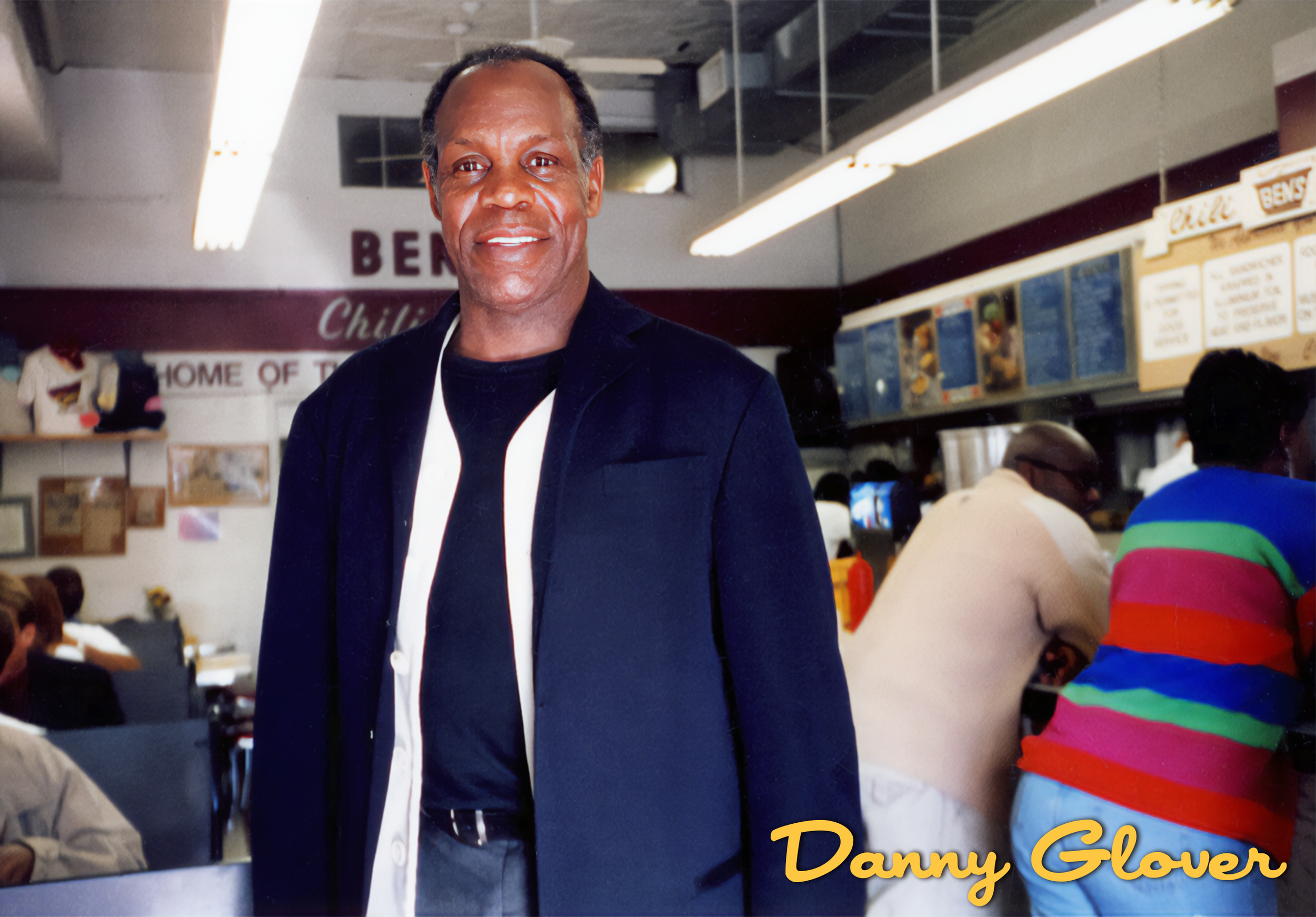 A man in a dark blazer and black shirt stands in a fast-food restaurant, smiling at the camera, with people in colorful clothing in the background. The restaurant has illuminated menu boards and signs, and the name 'Ben's' visible in the background. 
