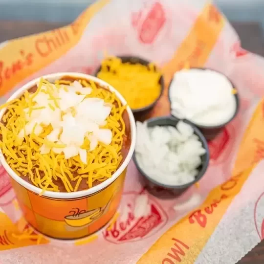 A Taco Bell meal with a cup of chilli topped with shredded cheese and diced onions, accompanied by three small containers of sour cream, guacamole, and diced onions.